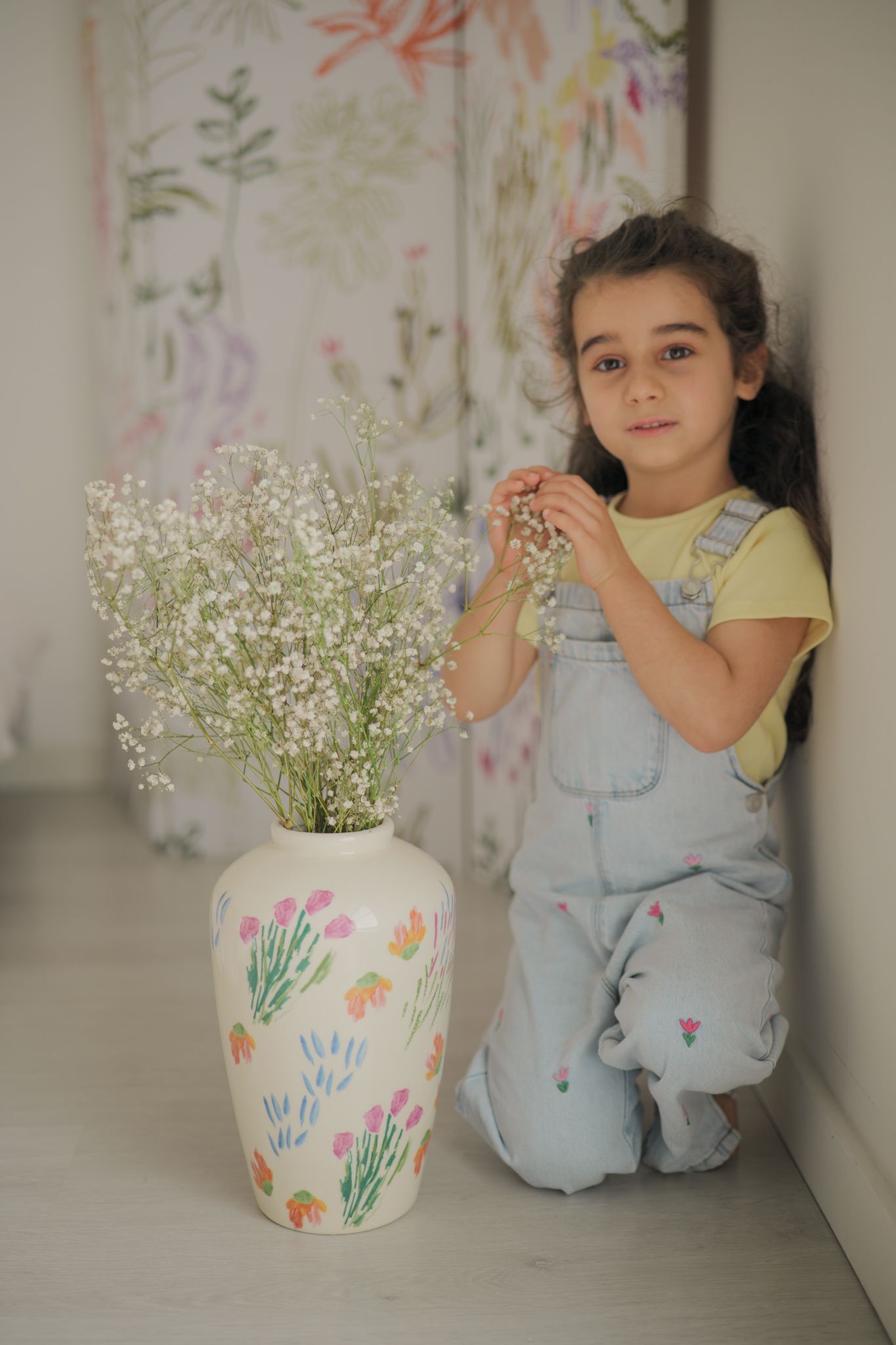 Young girl in denim overalls kneeling beside a floral vase with white baby's breath flowers against a floral backdrop.