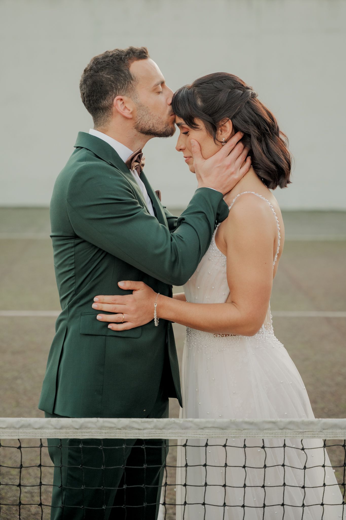 A couple in wedding attire shares a romantic moment by a tennis net on their special day.
