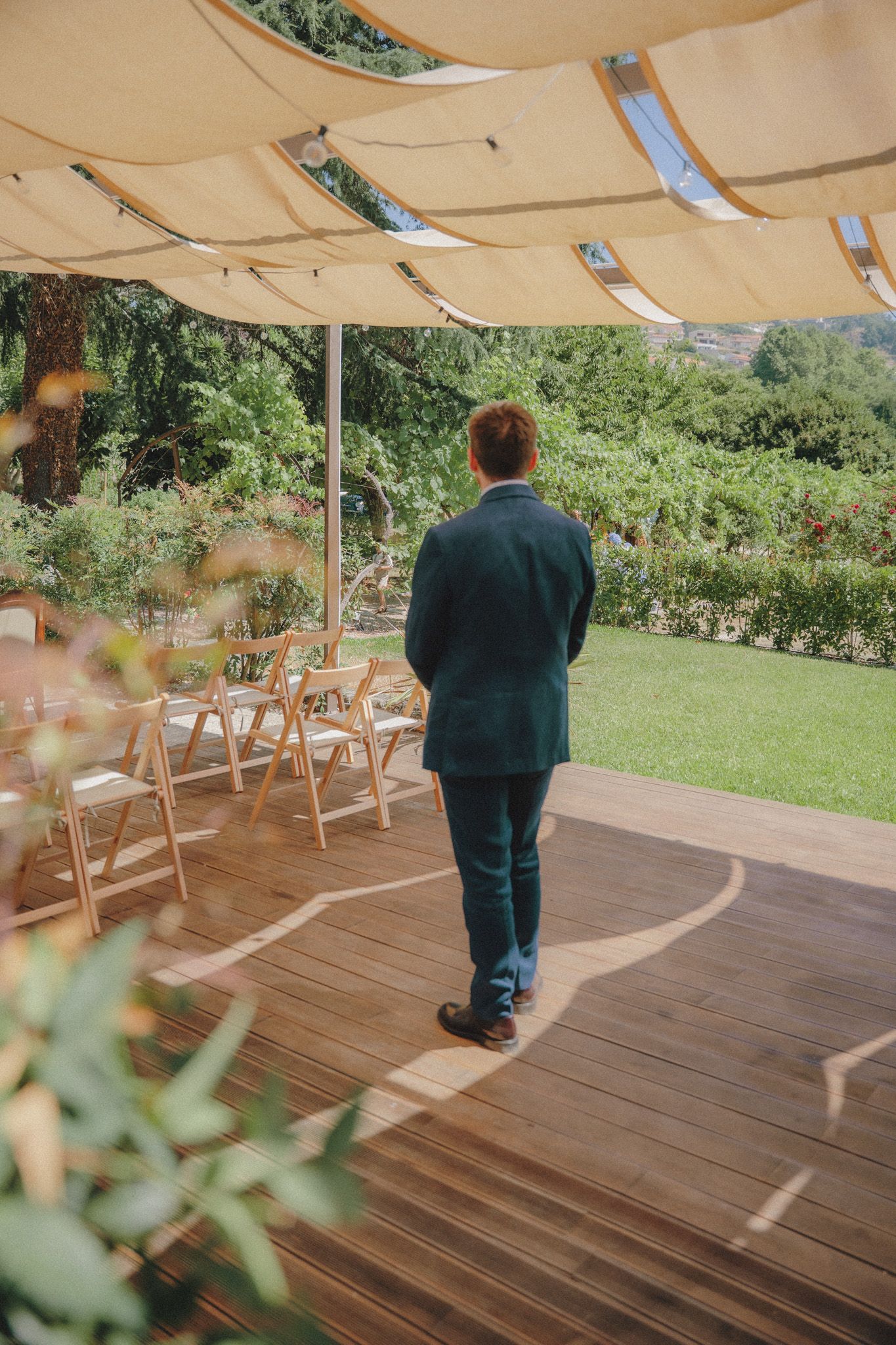 Person in formal attire walks across a wooden deck under a beige canopy with outdoor seating and greenery in background.