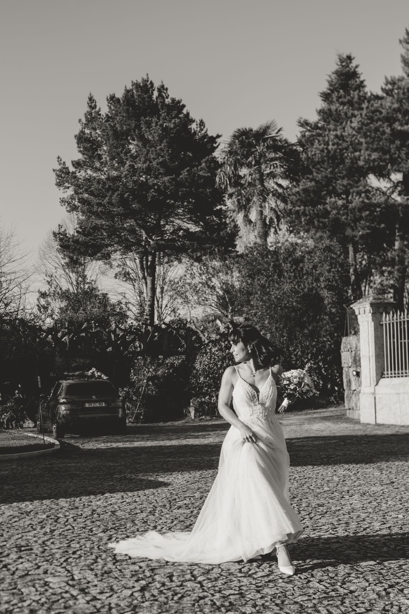 A bride in a flowing white gown poses outdoors near pine trees and a white gate.