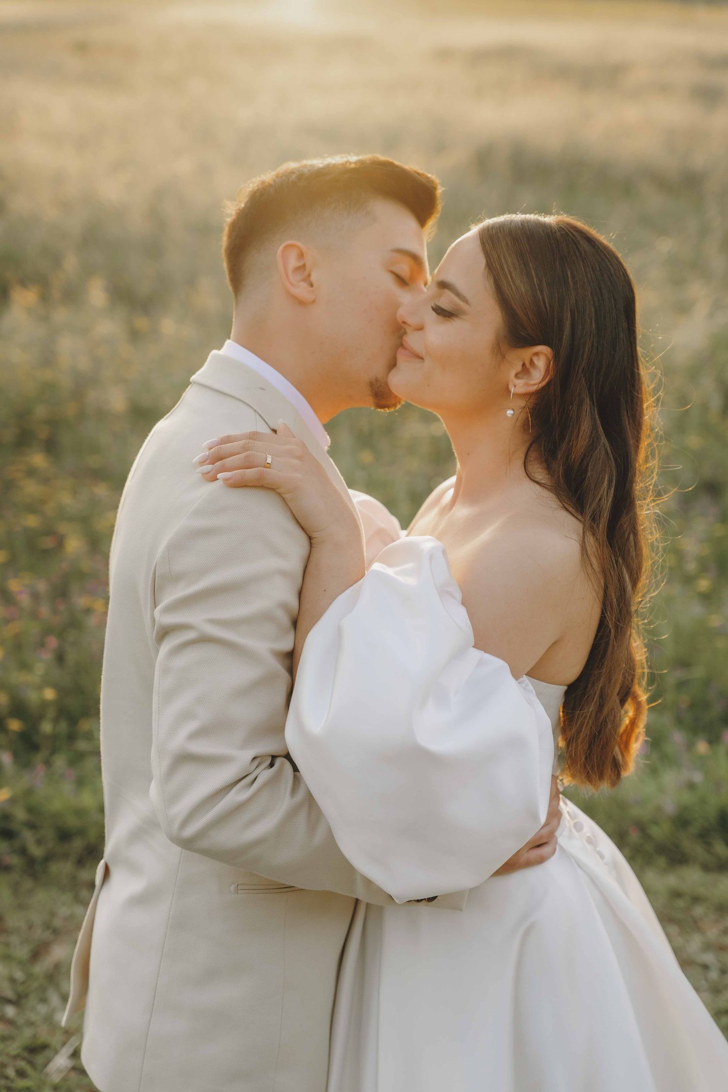 A couple in wedding attire share tender kisses in a sunlit field during golden hour.