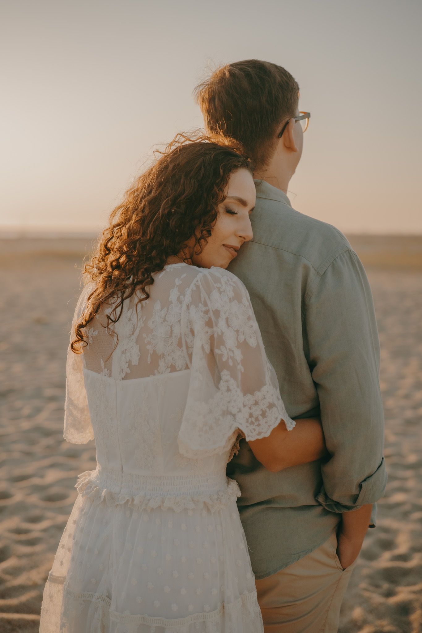 A romantic couple in wedding attire embraces on a sandy beach at sunset.