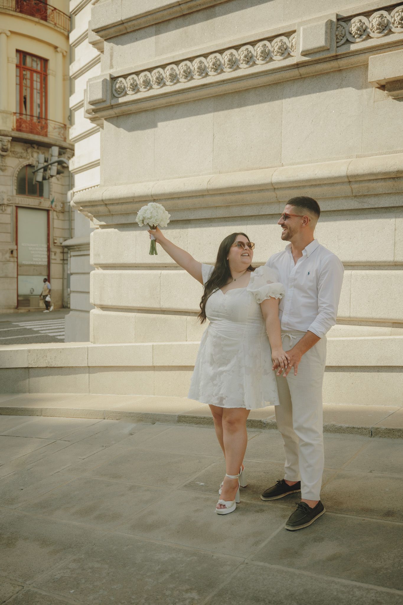 A romantic couple in white clothing poses against a grand building exterior.