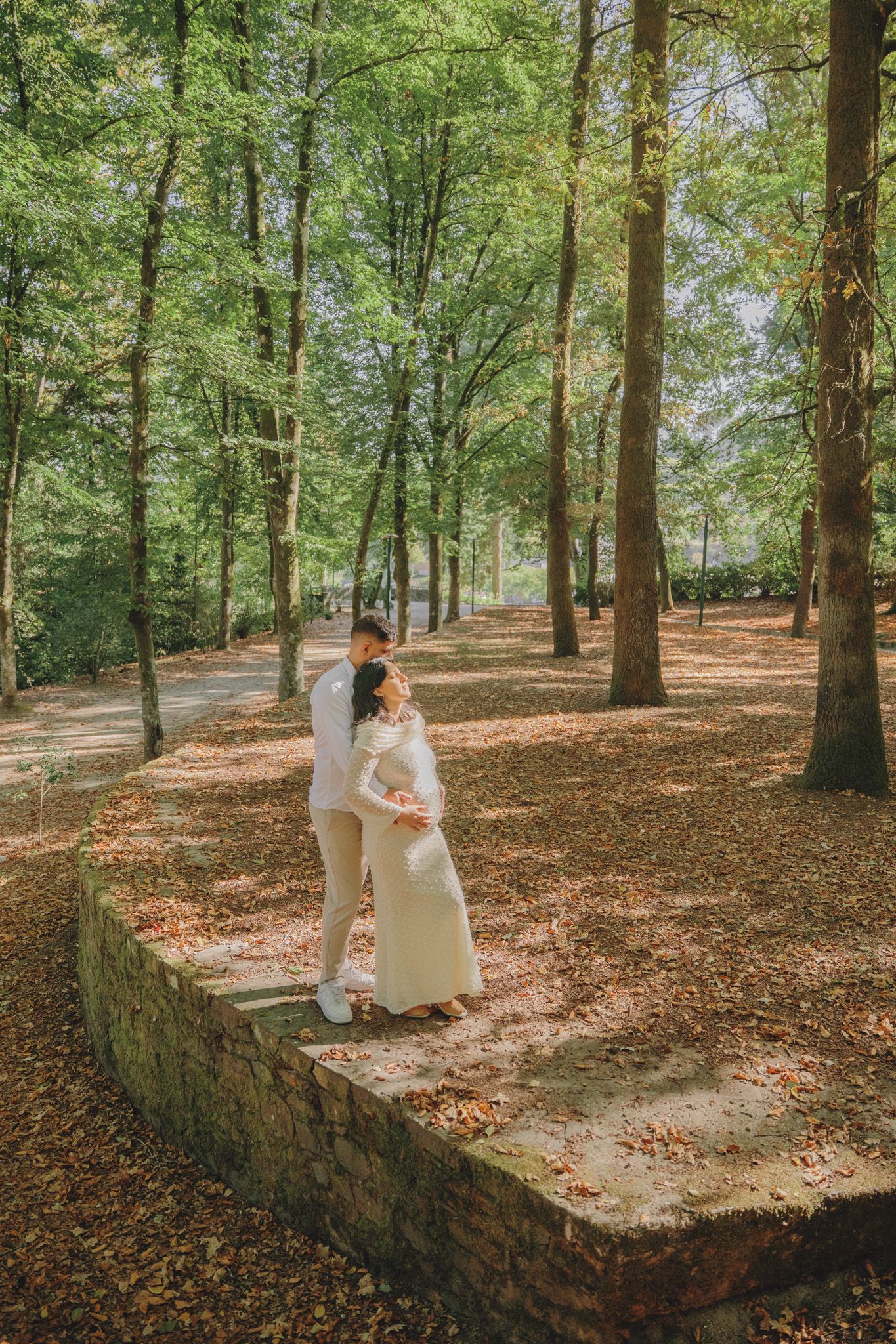 Person in white dress walks along stone wall in autumn forest.