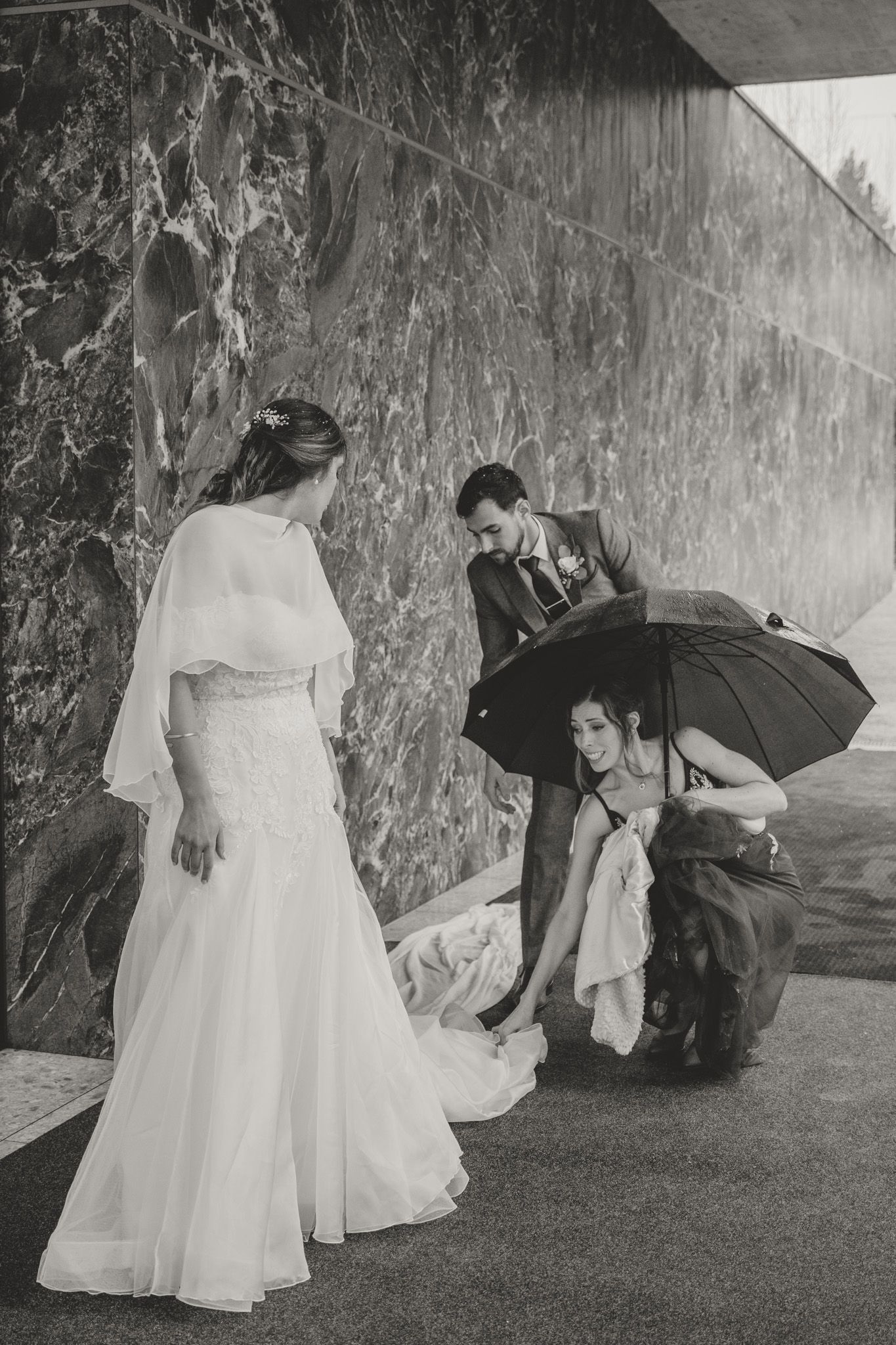 Wedding party with umbrellas navigating rainy weather on grassy slope.