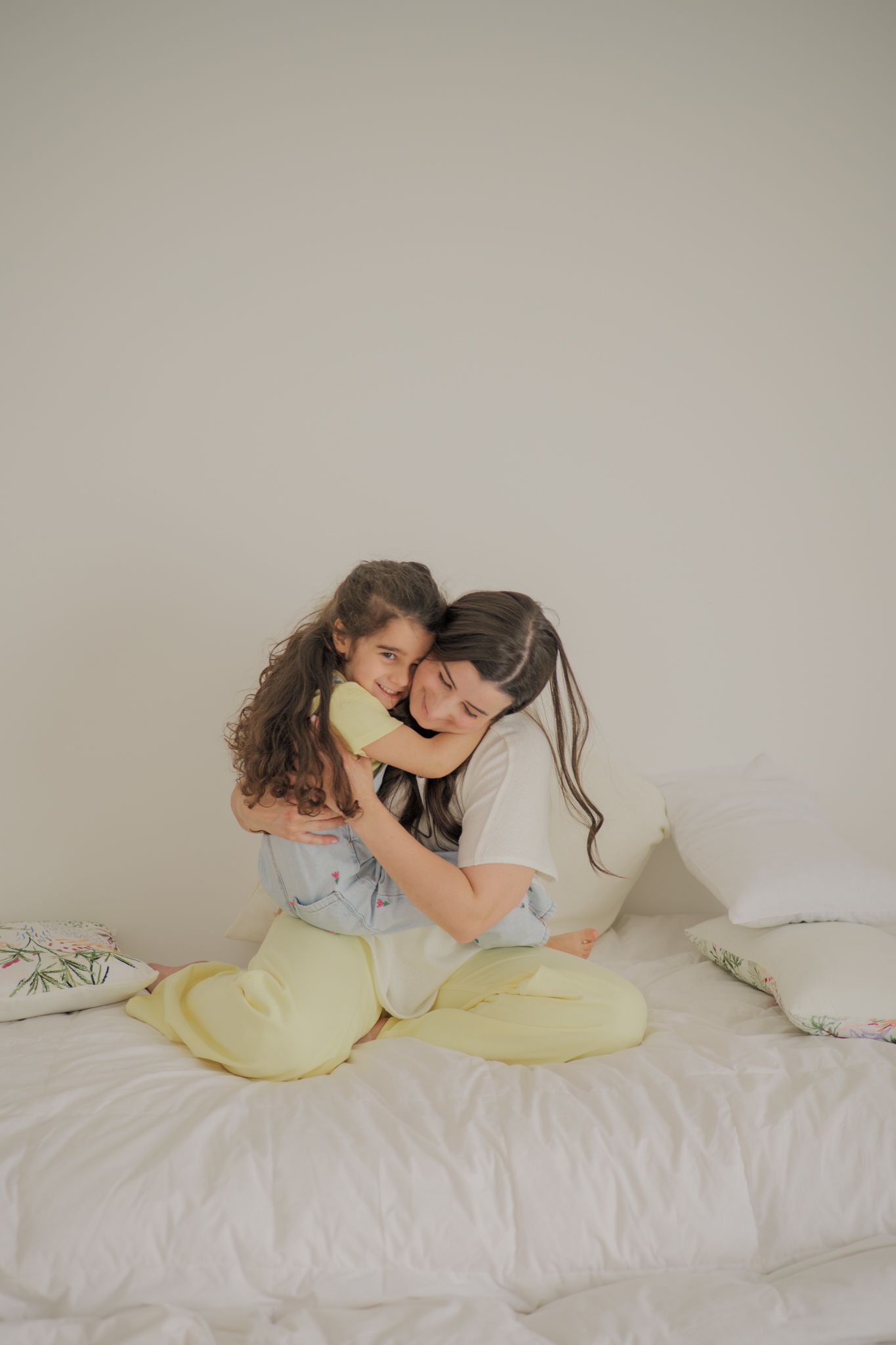 A mother and young daughter share a warm hug while sitting on a white bed, both wearing pastel pajamas.