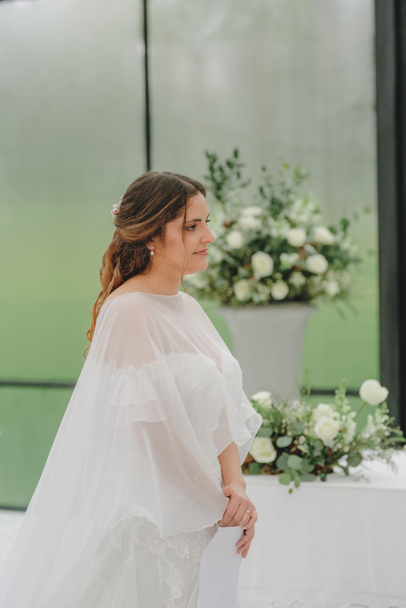 Bride in flowing white dress poses near elegant floral arrangements in a greenhouse setting.