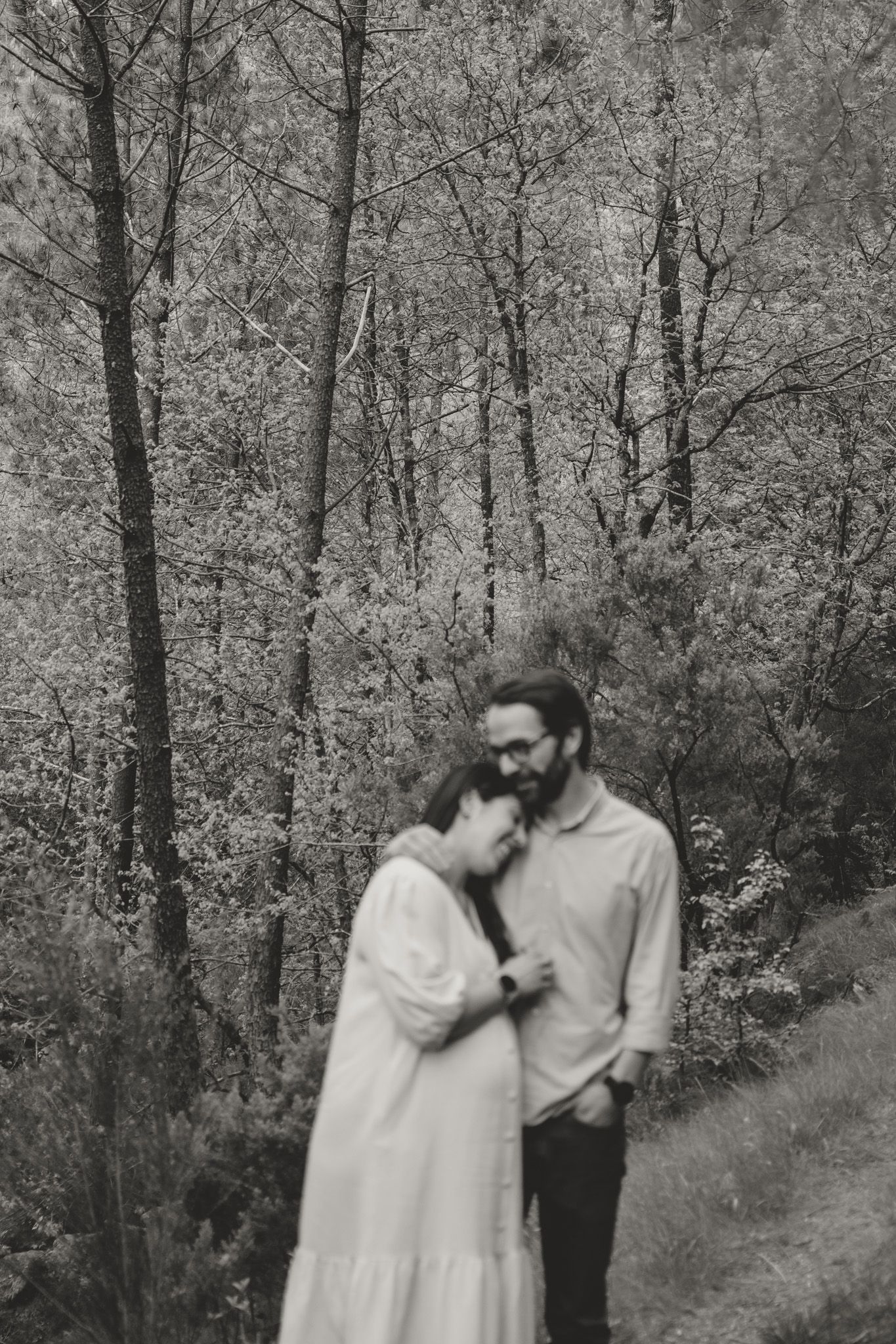 Black and white photograph of a couple embracing on a wooded path surrounded by trees.