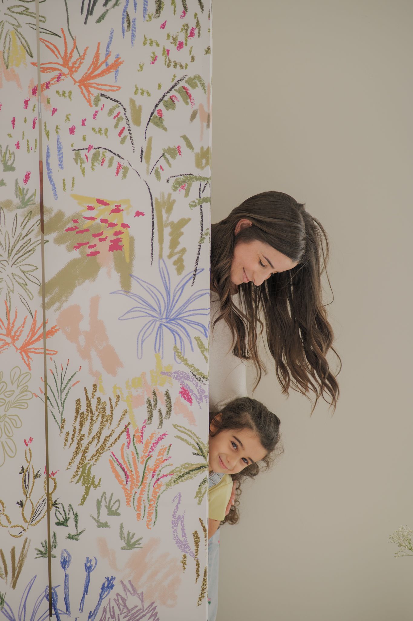 Two women peek playfully around a colorful floral wallpapered wall, smiling warmly in a bright, cheerful room.