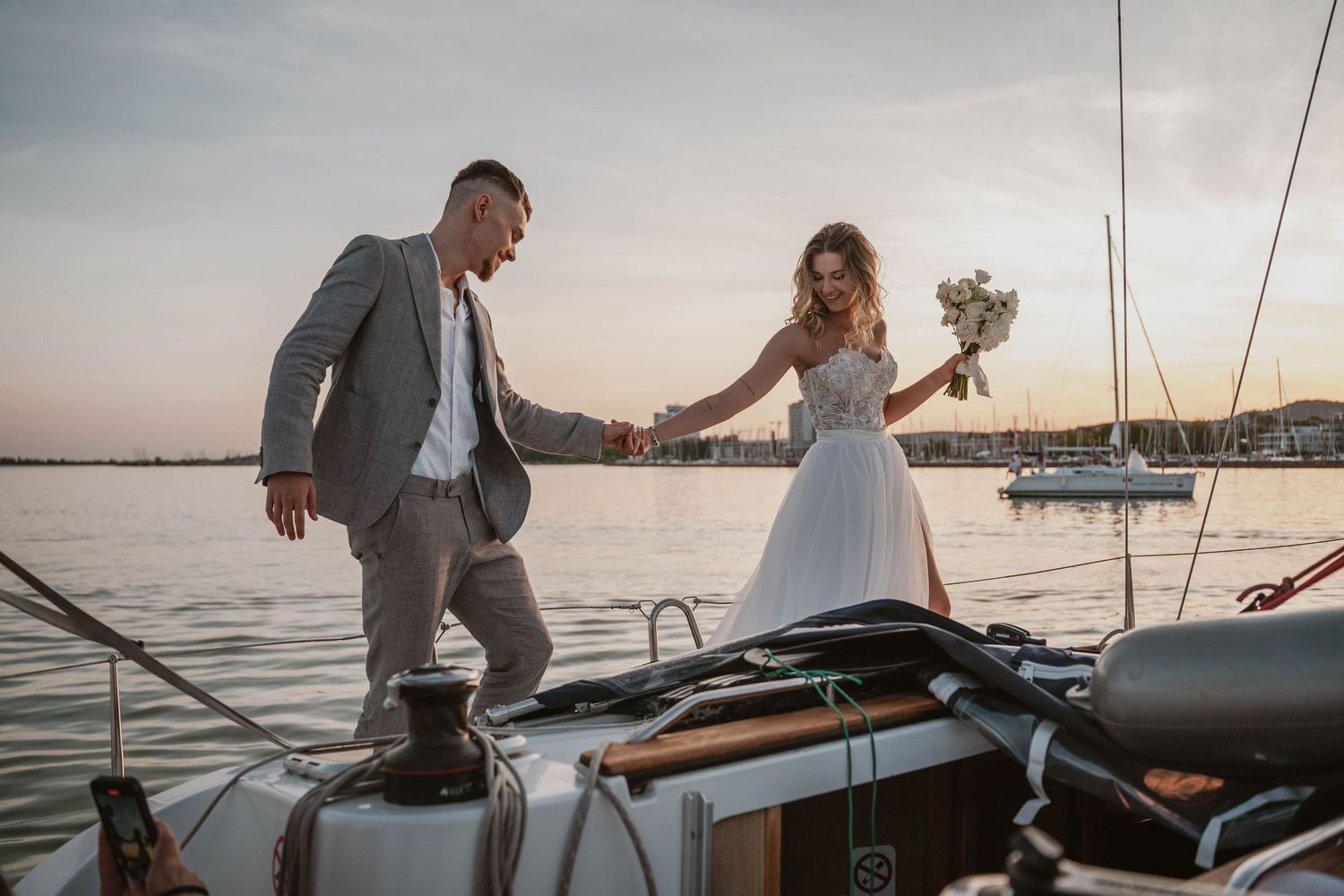 Wedding couple stands together holding hands on a sailboat deck at sunset with marina in the background.
