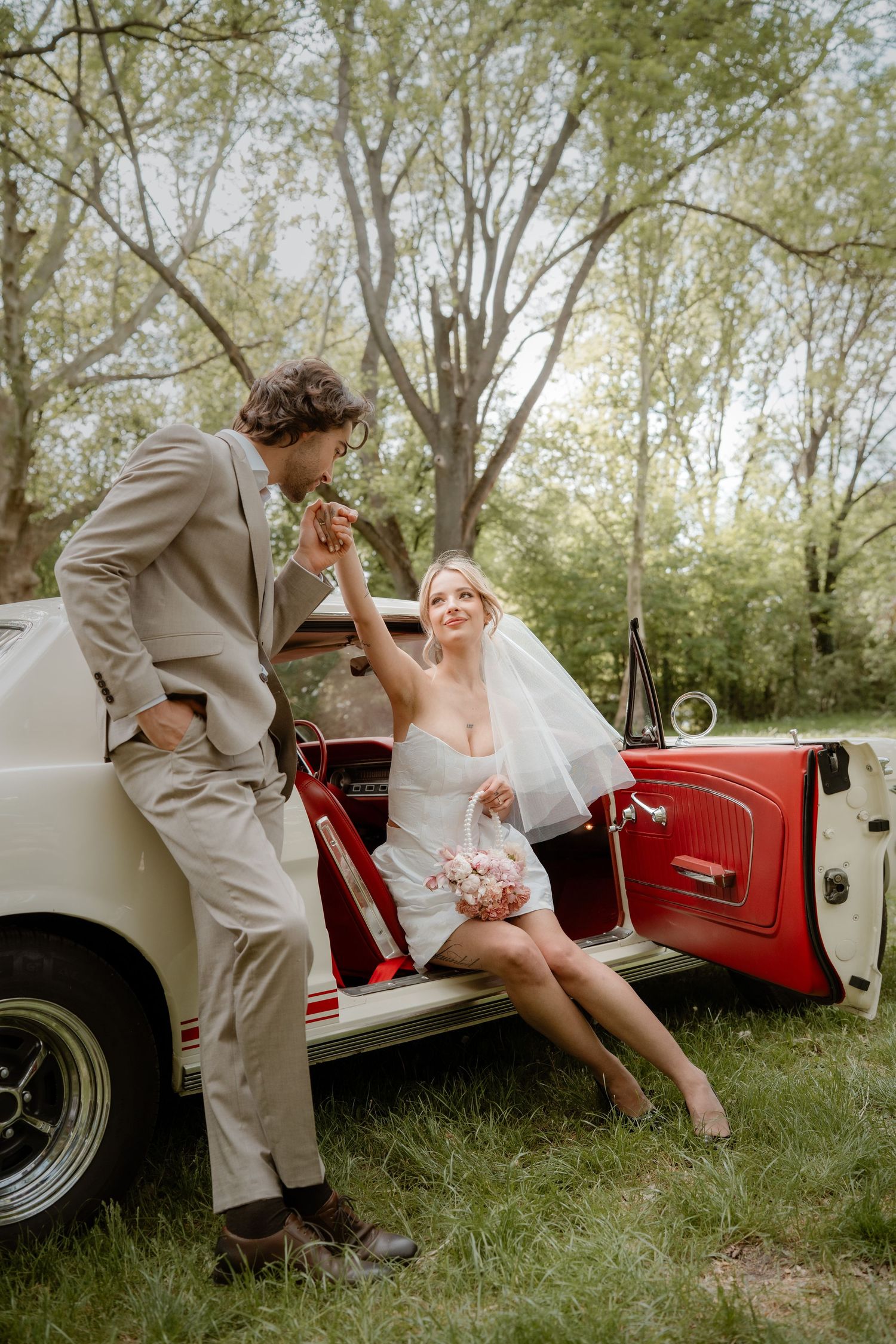 Bride in white mini dress sits in red vintage car as groom in tan suit kisses her hand outdoors.