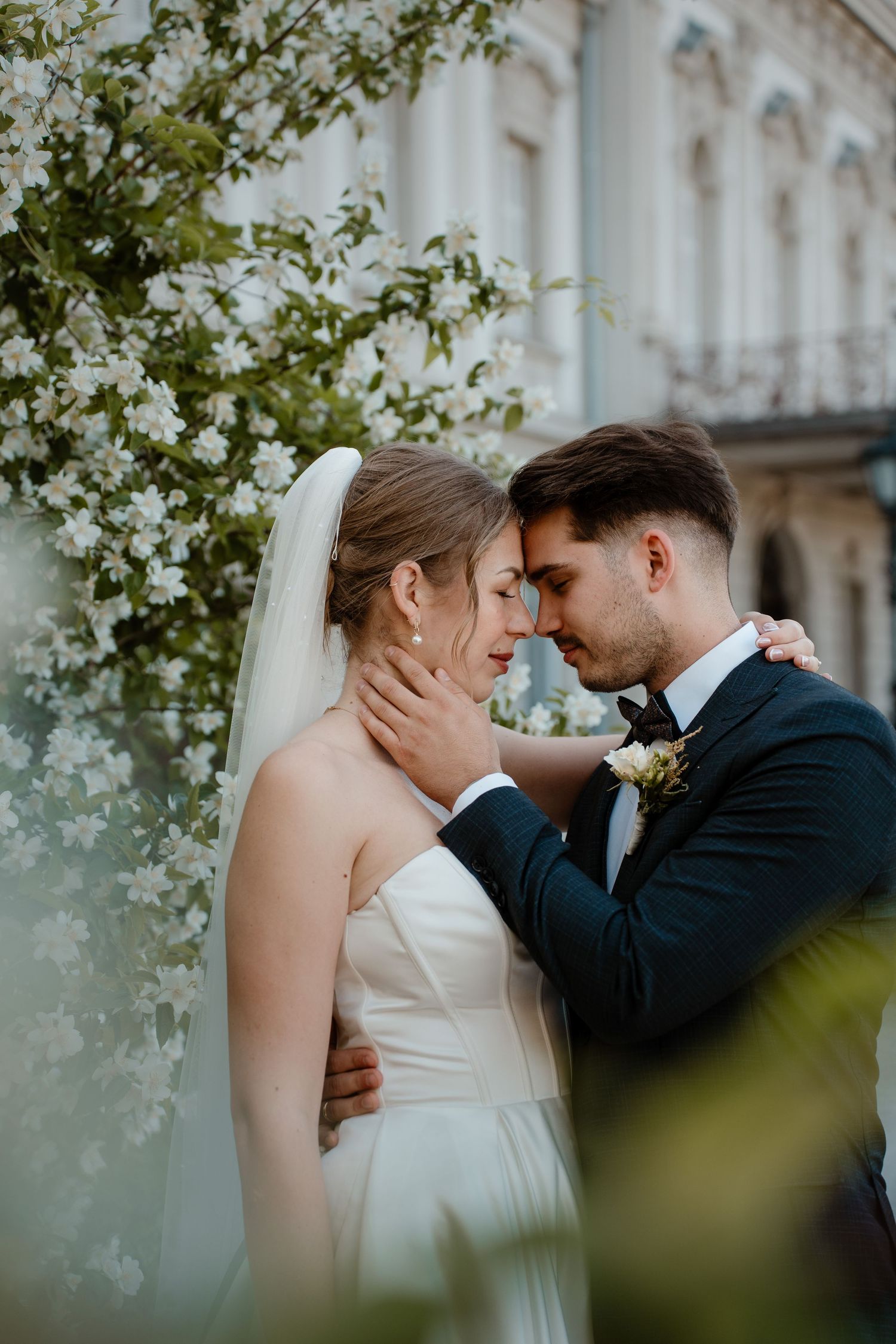 A bride and groom share an intimate moment, foreheads touching, near white blossoms outside an elegant building.