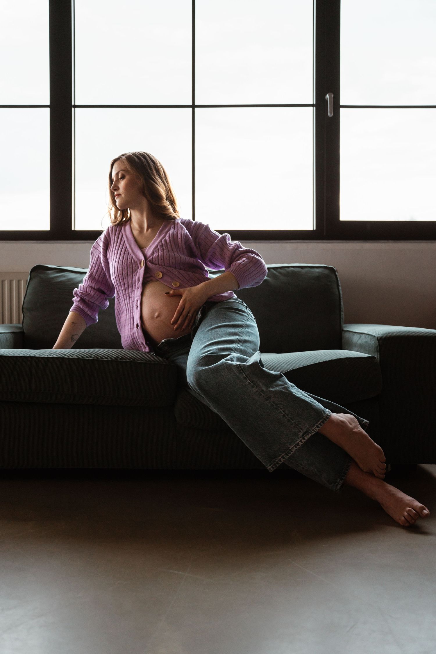 A relaxed figure lounges on a gray couch by large windows wearing a lavender sweater and cropped jeans.