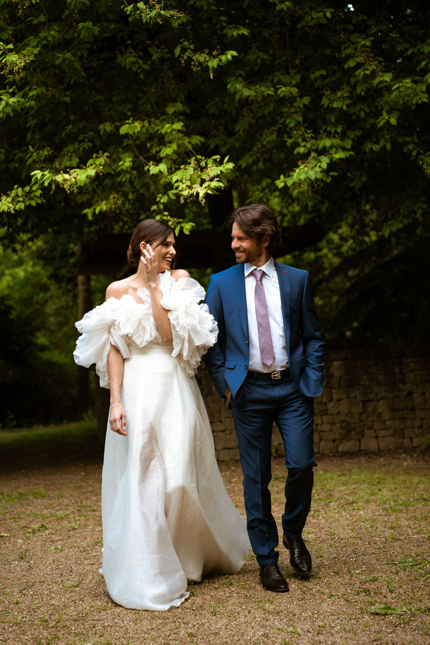 A couple in elegant wedding attire walk together on a garden path with lush green trees in the background.