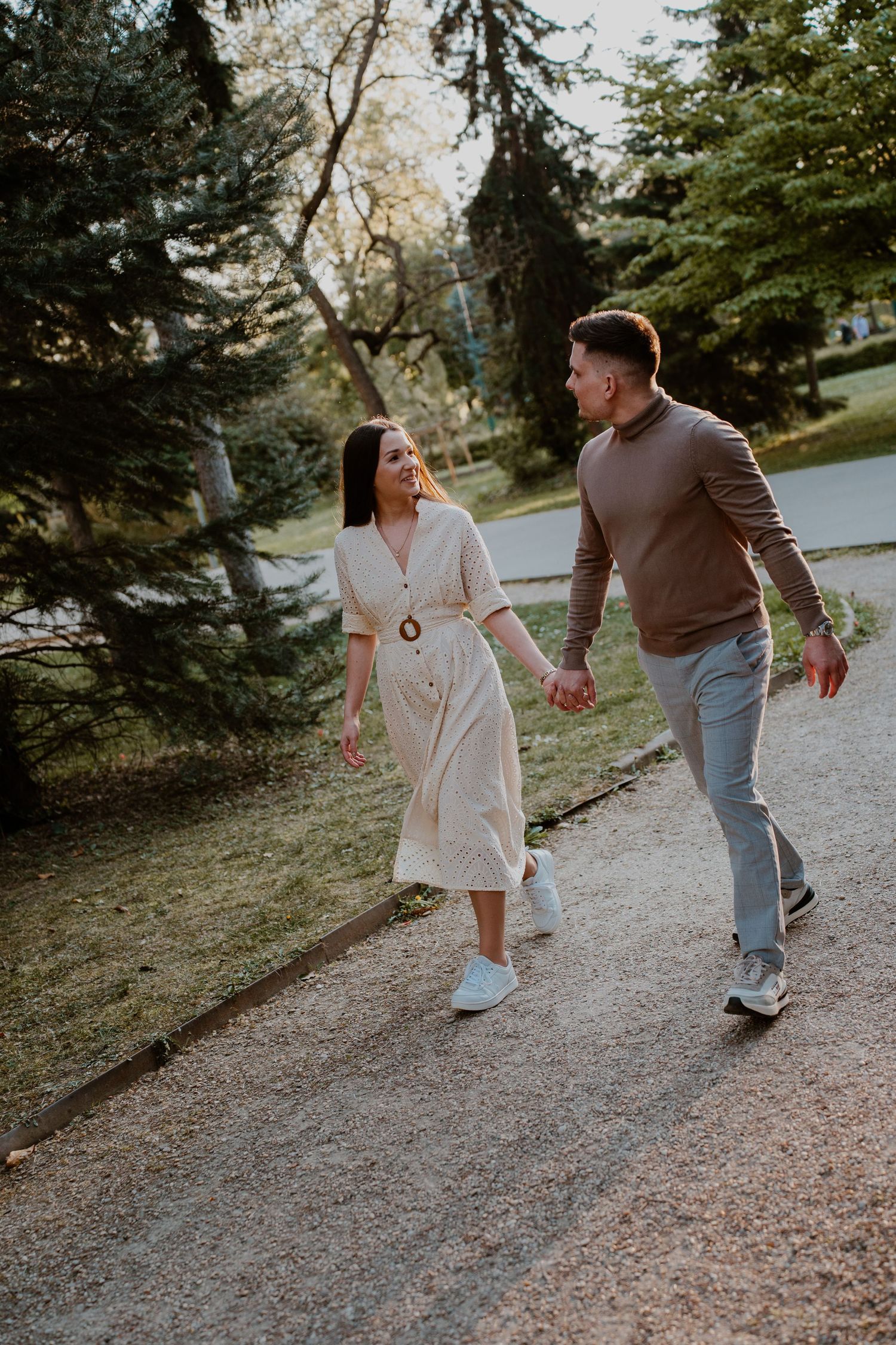 Happy couple holding hands while walking together on a park path surrounded by lush green trees.