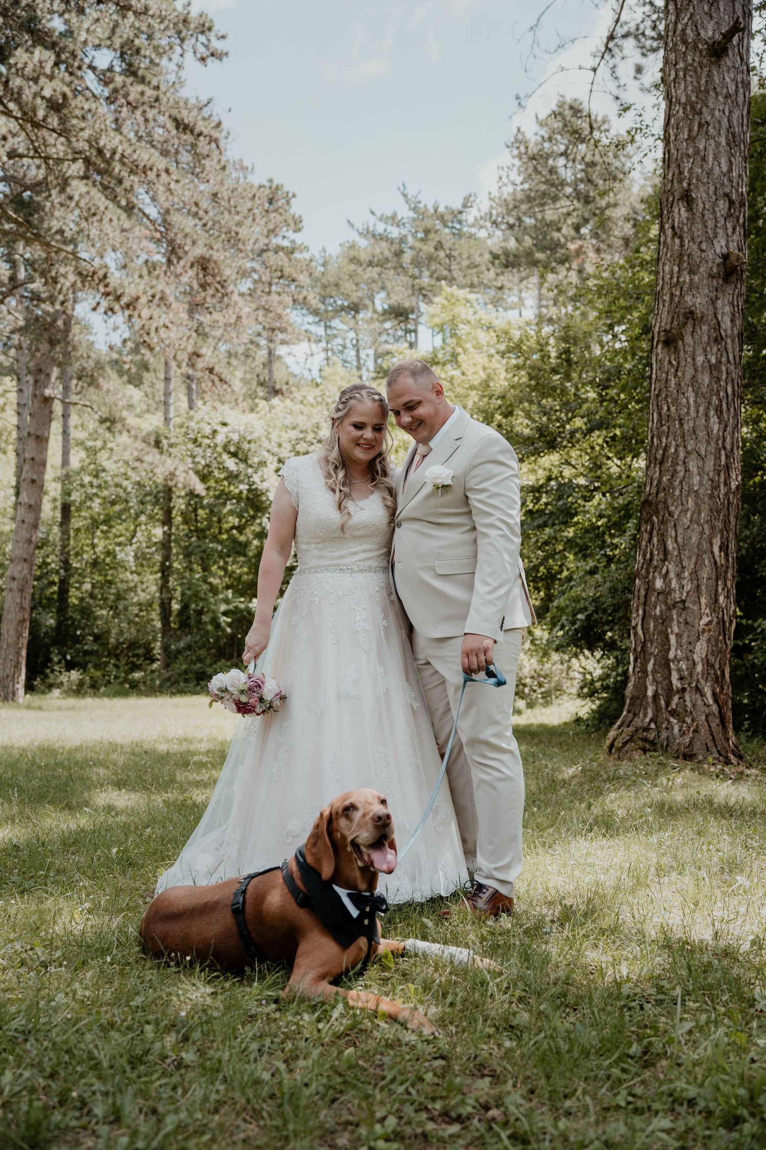 Bride and groom posing with their Bloodhound dog wearing a bow tie in a lush green forest setting on their wedding day.