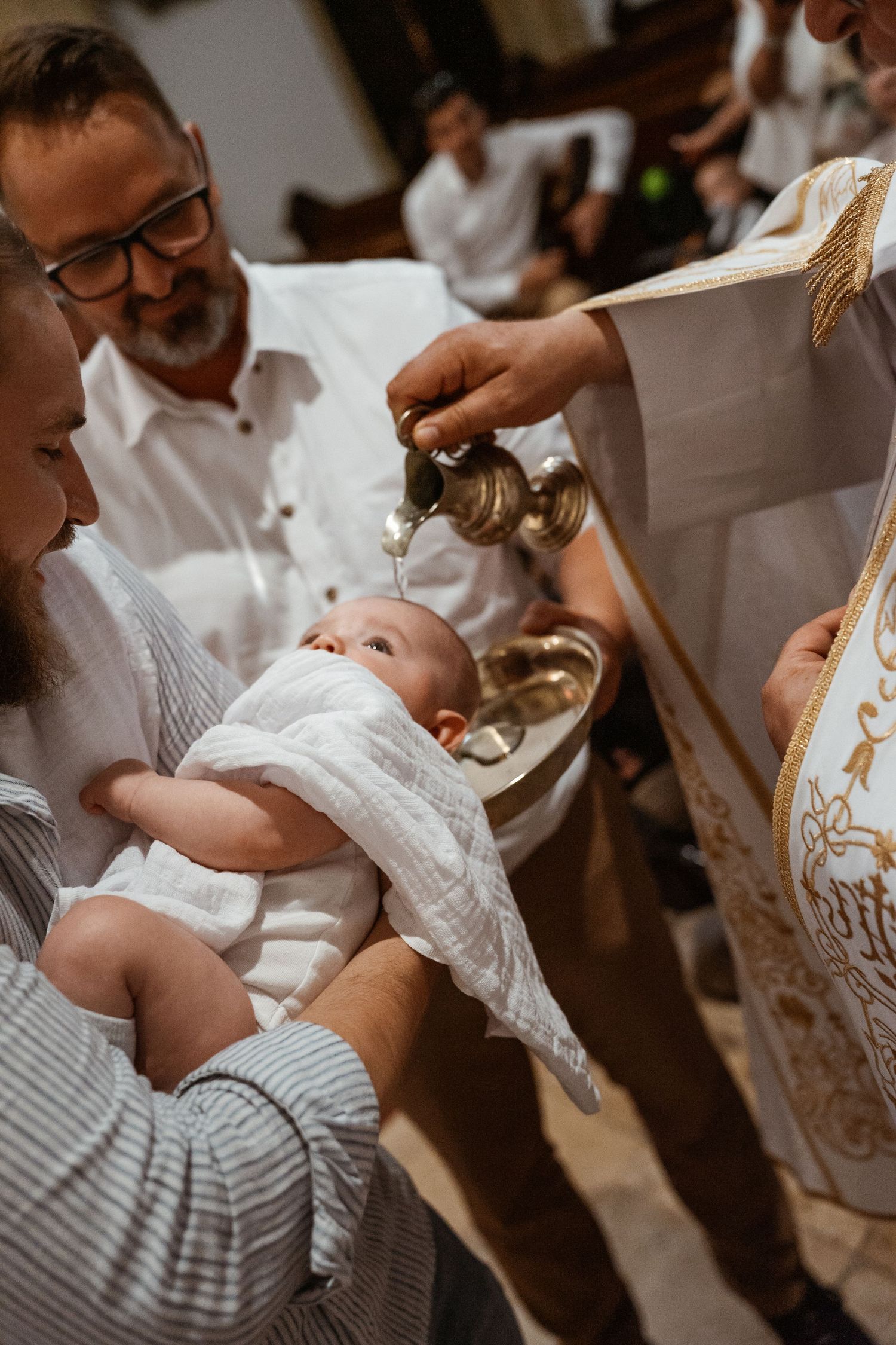 A baby in white christening gown receives holy water during a traditional baptism ceremony.