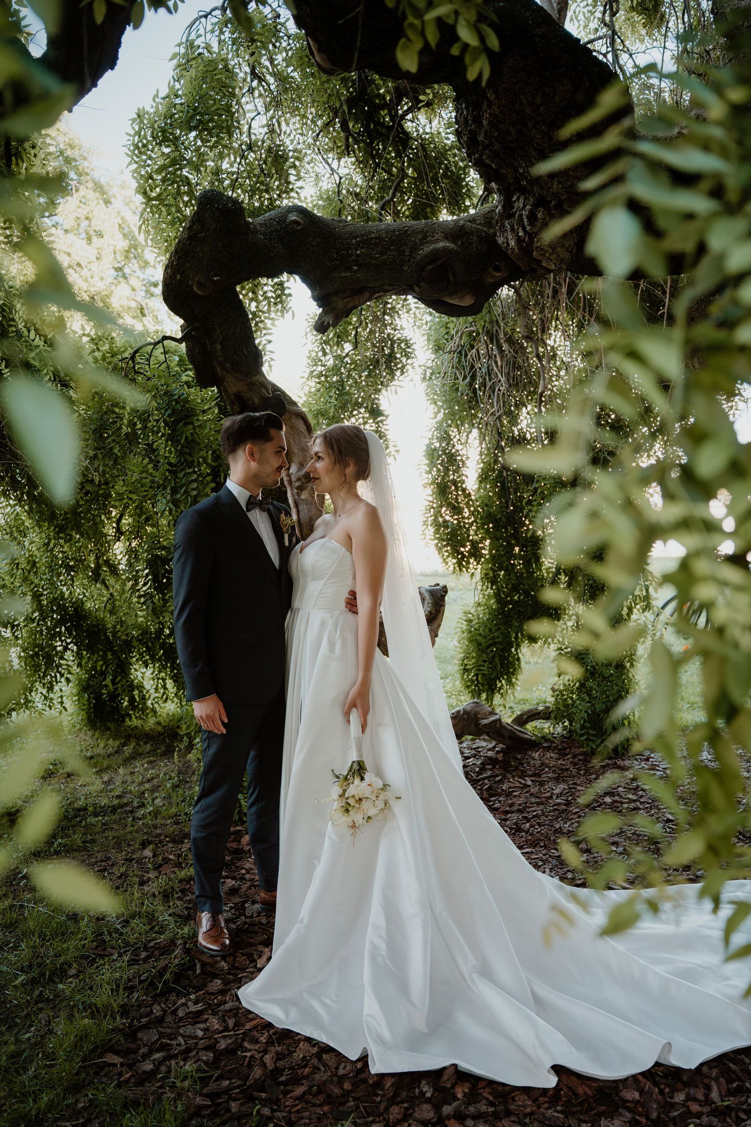 Bride and groom share a romantic moment under a large tree, surrounded by lush greenery, as she holds a white bouquet.