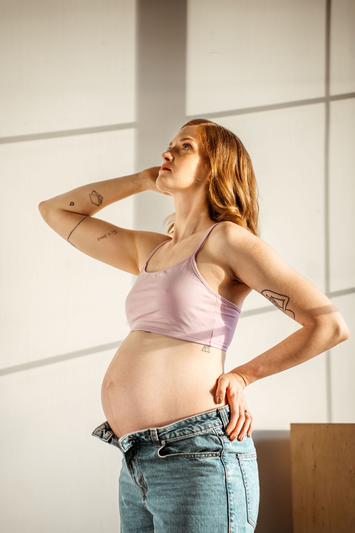 Expectant person in pink crop top and jeans stands in sunlit room showing pregnancy bump against white wall.