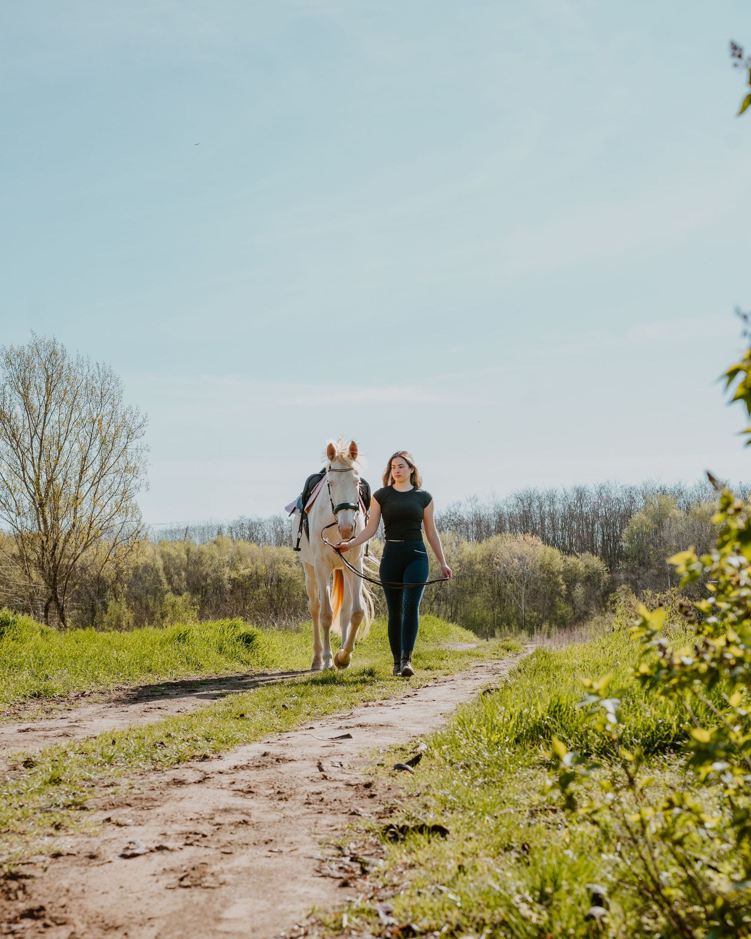 Person in black outfit walking with white horse along a scenic dirt path surrounded by spring greenery.