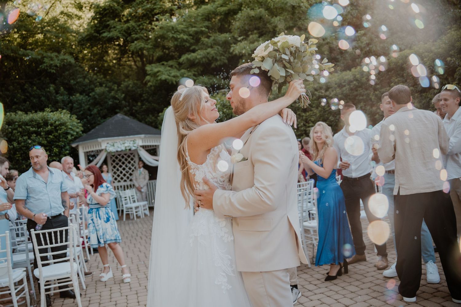 Bride and groom share first kiss outdoors at wedding reception with guests and colorful bubbles floating in background.