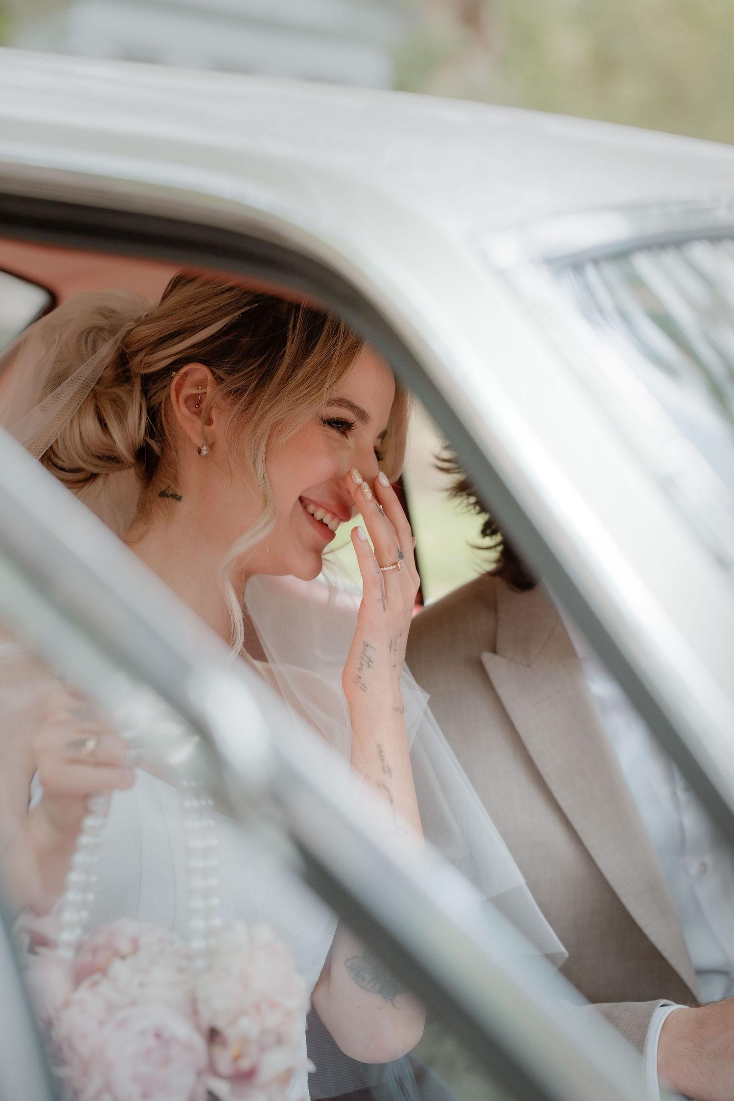 Smiling bride in wedding dress sitting in a car, covering her mouth with her hand, wearing a ring.
