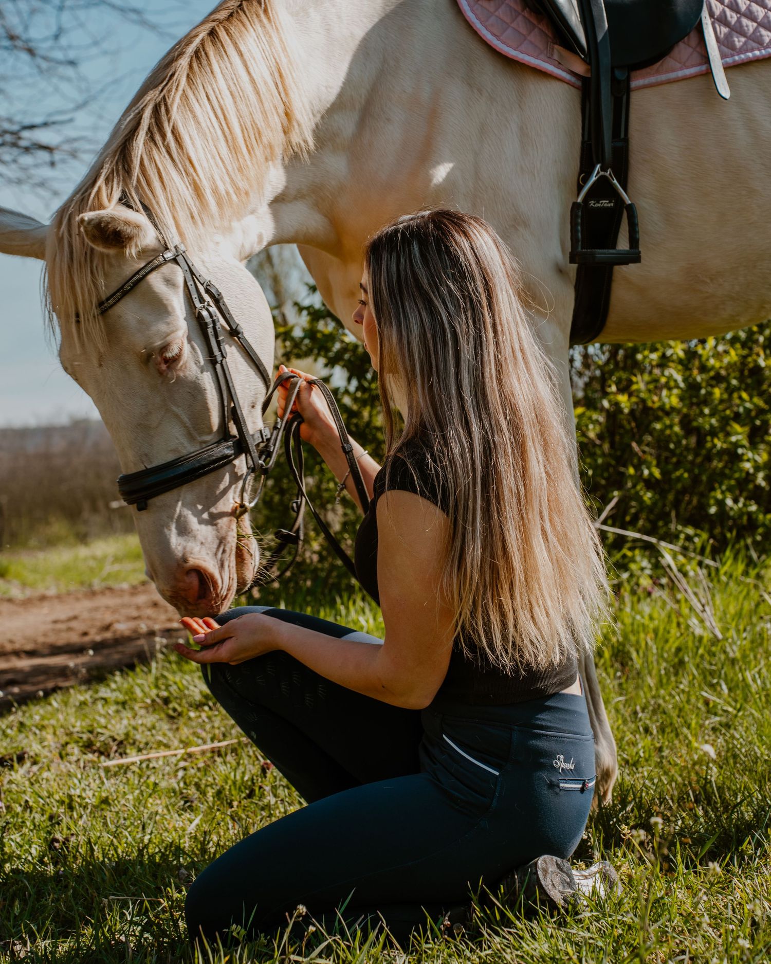 Long-haired person in black attire gently interacts with a white horse on a sunny day in a grassy outdoor setting.