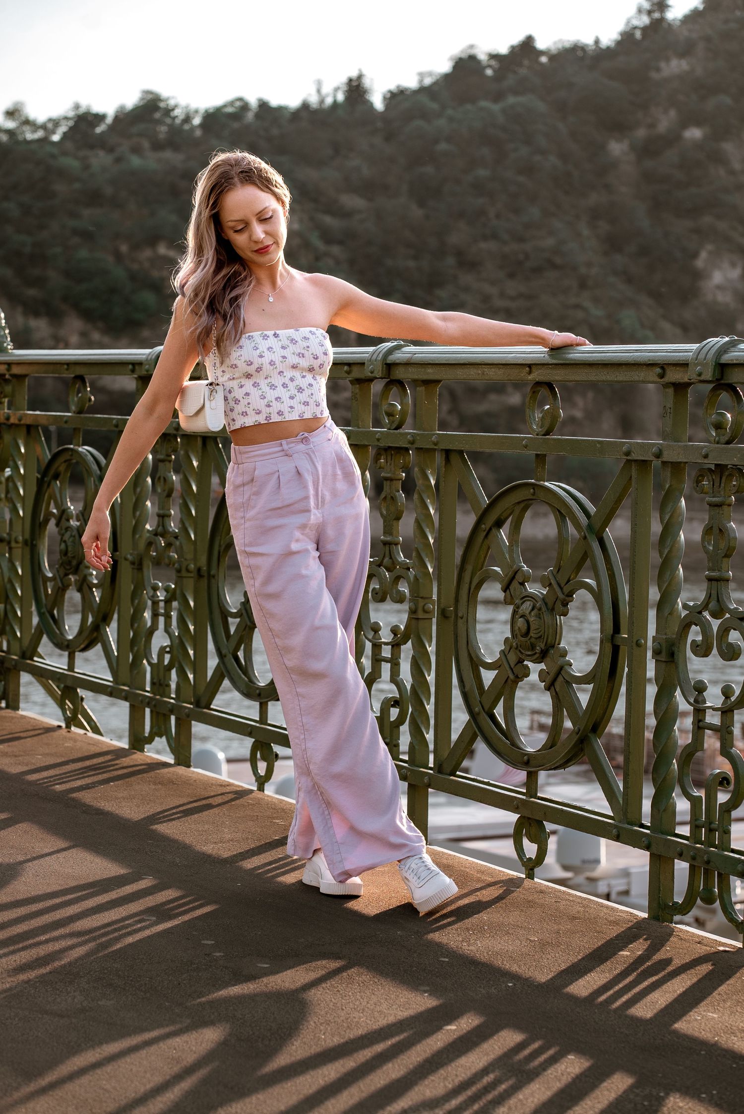 A person in a polka dot crop top and lavender pants stands gracefully on an ornate bridge with mountain views at sunset.