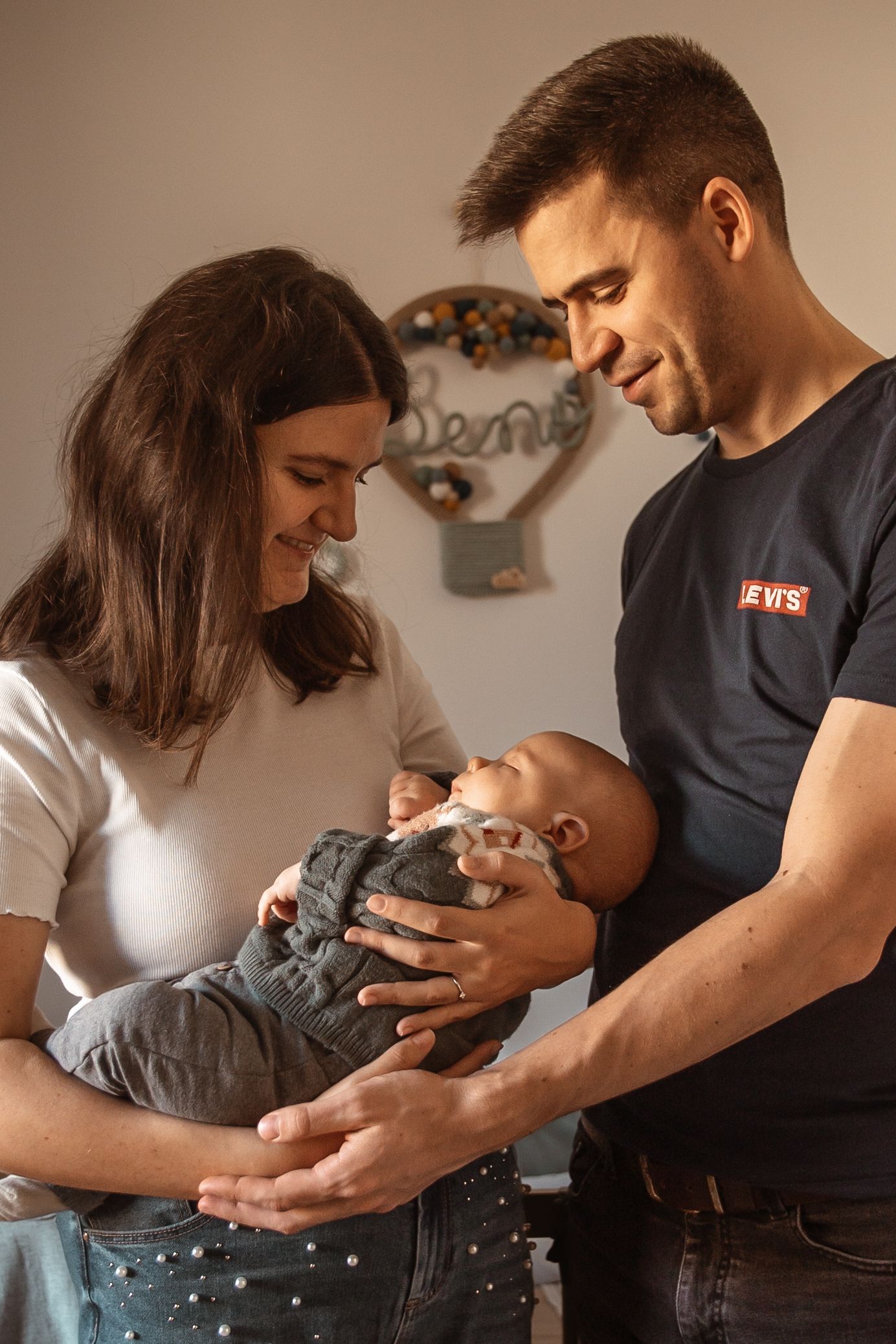 A couple in casual attire smiling while holding and looking down at their newborn baby in a cozy home setting.