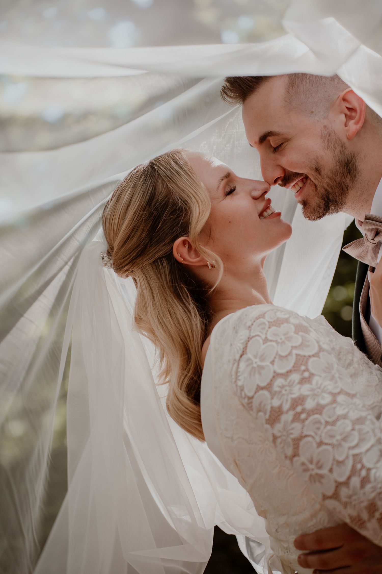 A bride and groom share a romantic almost-kiss under a flowing white veil on their wedding day.