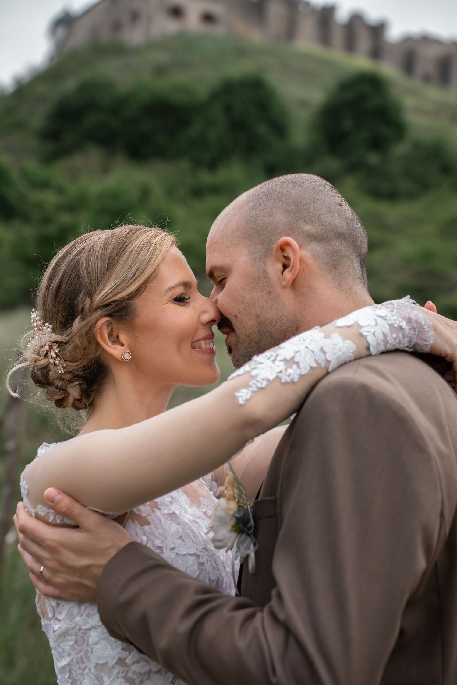 A romantic wedding moment captured at a hilltop castle venue, with bride in lace dress and groom in brown suit embracing.