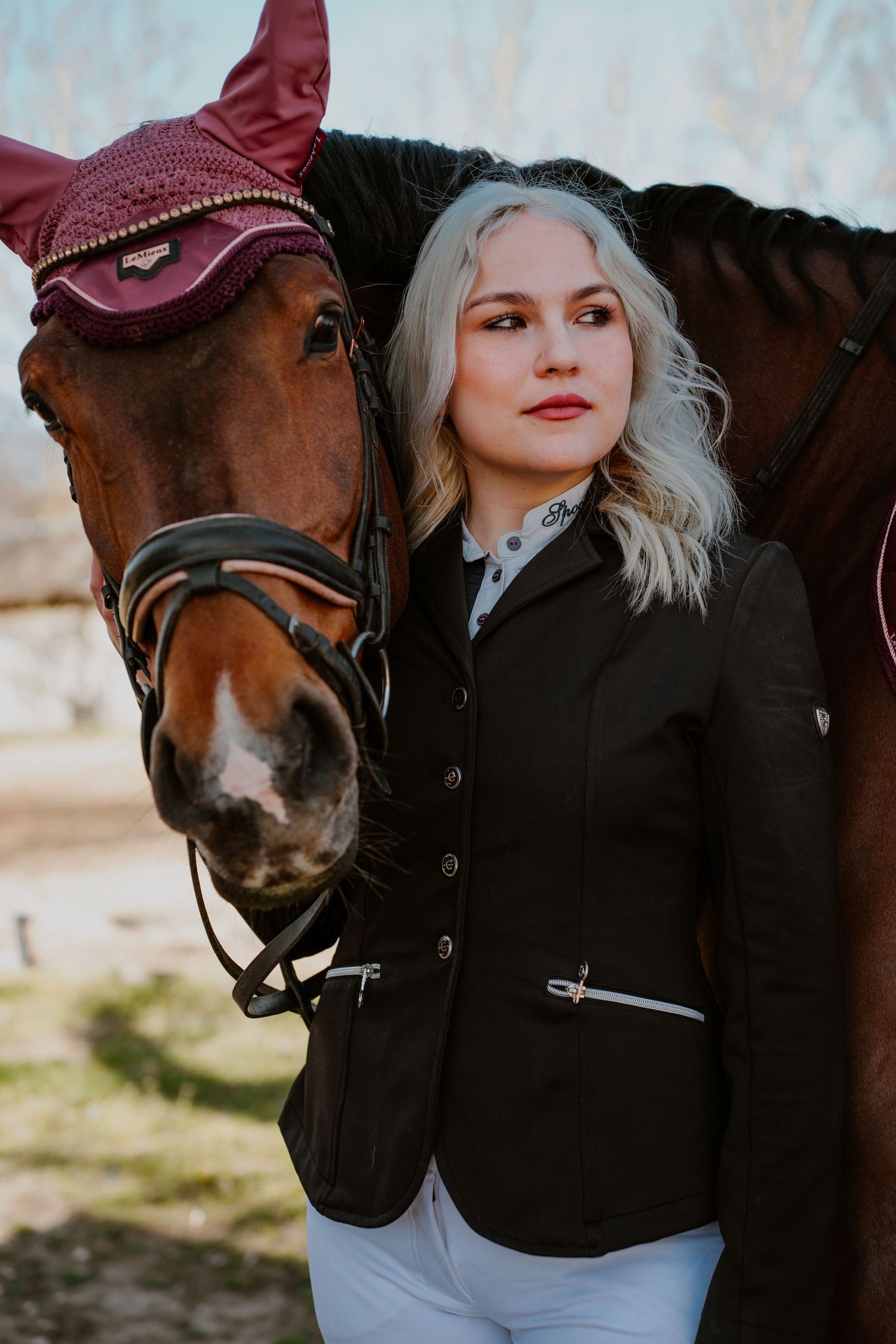 A portrait of an equestrian rider in a black blazer standing next to their dark brown horse with a burgundy ear bonnet.