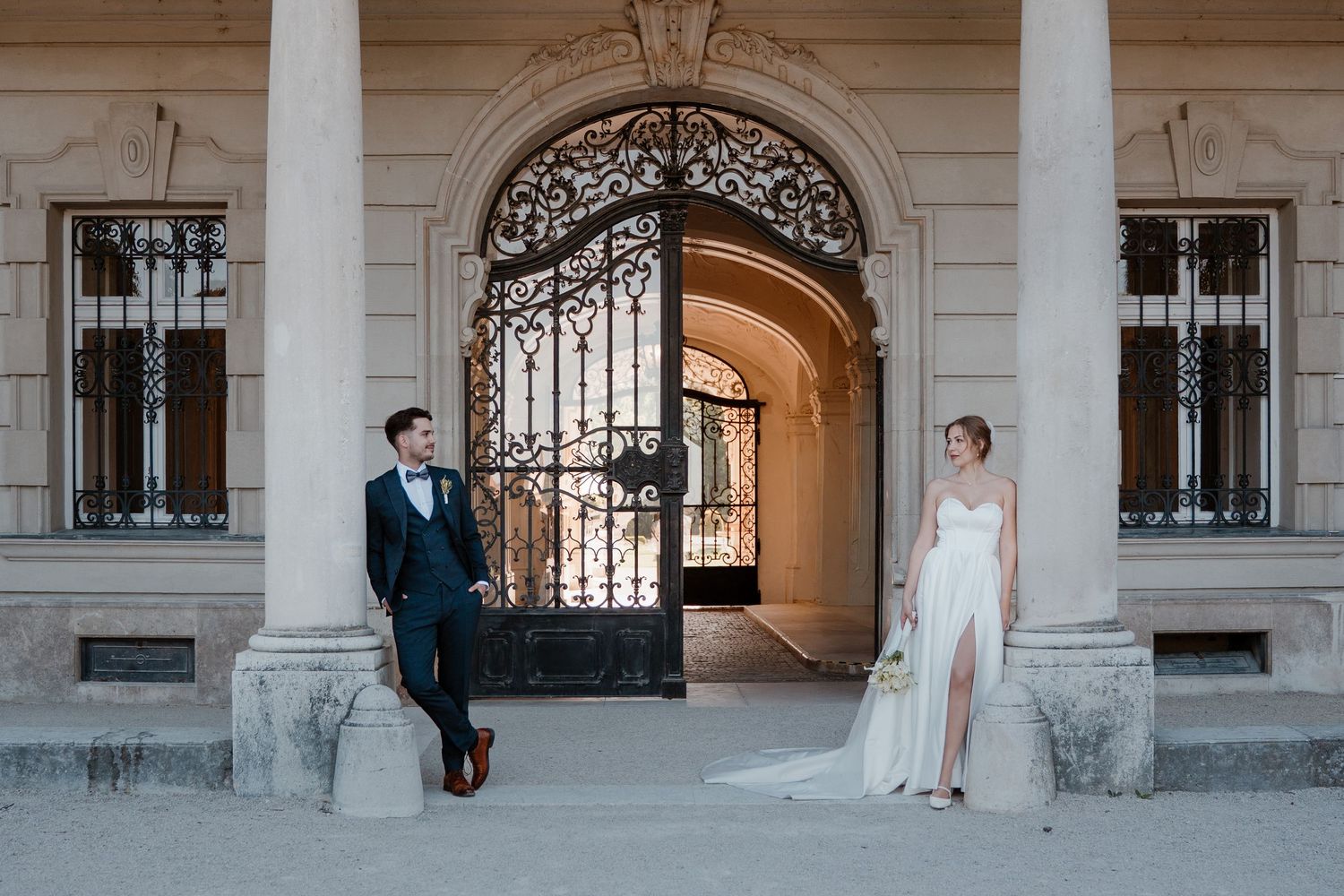 Bride and groom posing at ornate iron gate entrance of elegant European palace building with stone columns.