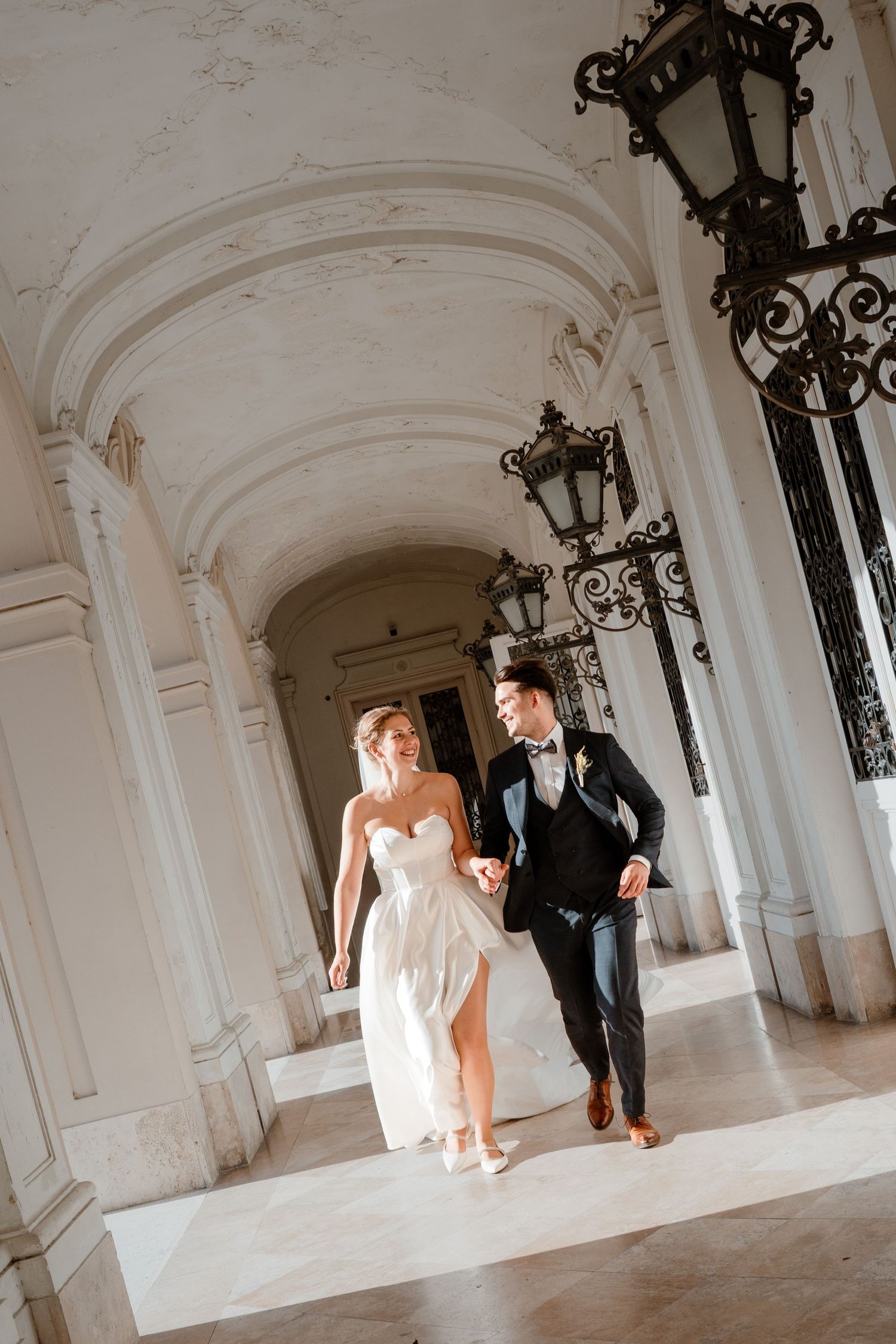 Bride and groom walk hand in hand through elegant white marble archway with ornate iron lanterns on their wedding day.