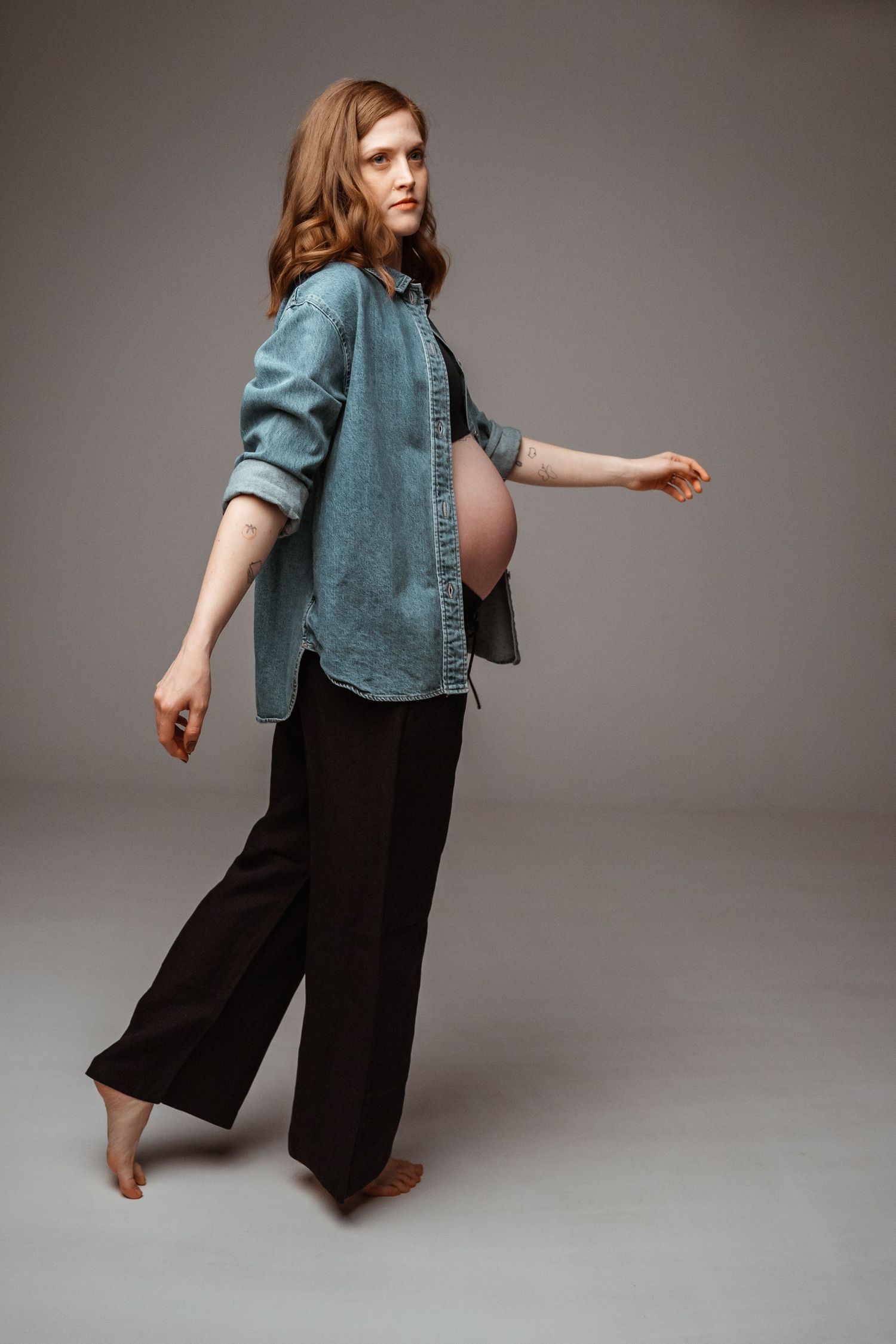 Person in denim button-up shirt and wide-leg black pants poses barefoot against grey studio backdrop.