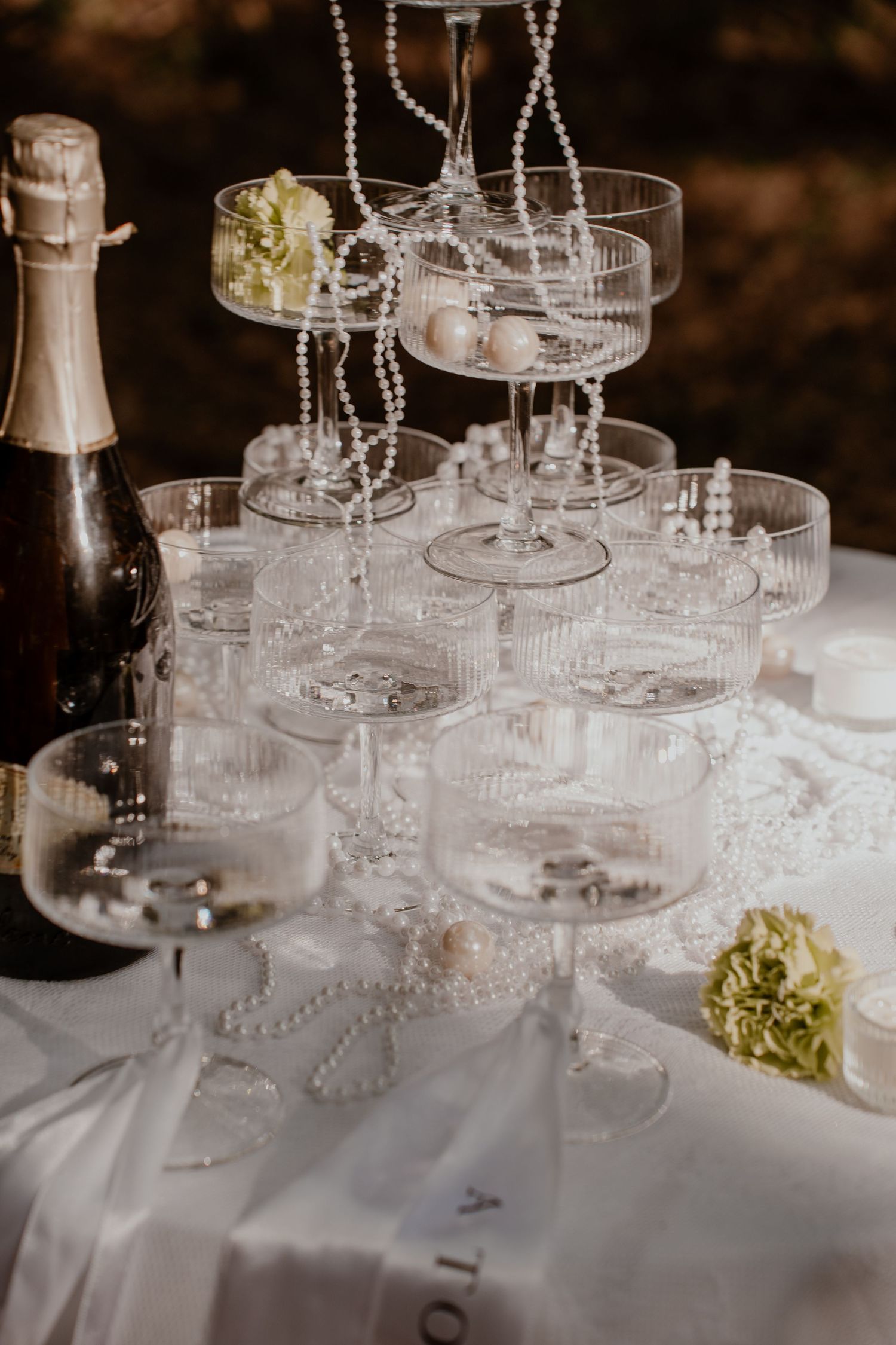 Elegant champagne tower with crystal coupe glasses, pearl strands, and a bottle of sparkling wine on a white tablecloth.