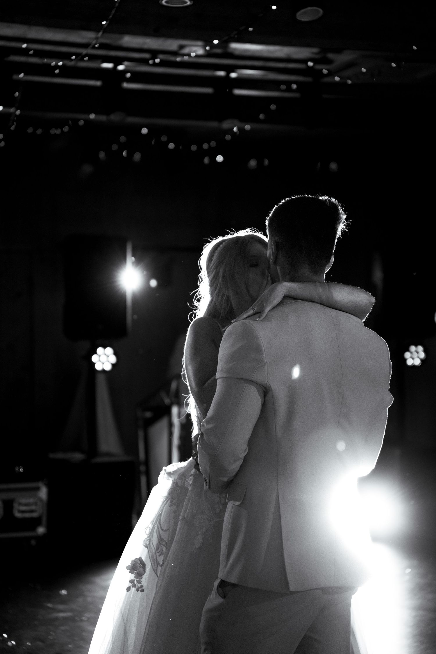 Black and white photo of bride and groom sharing first dance, backlit by stage lights at wedding reception.