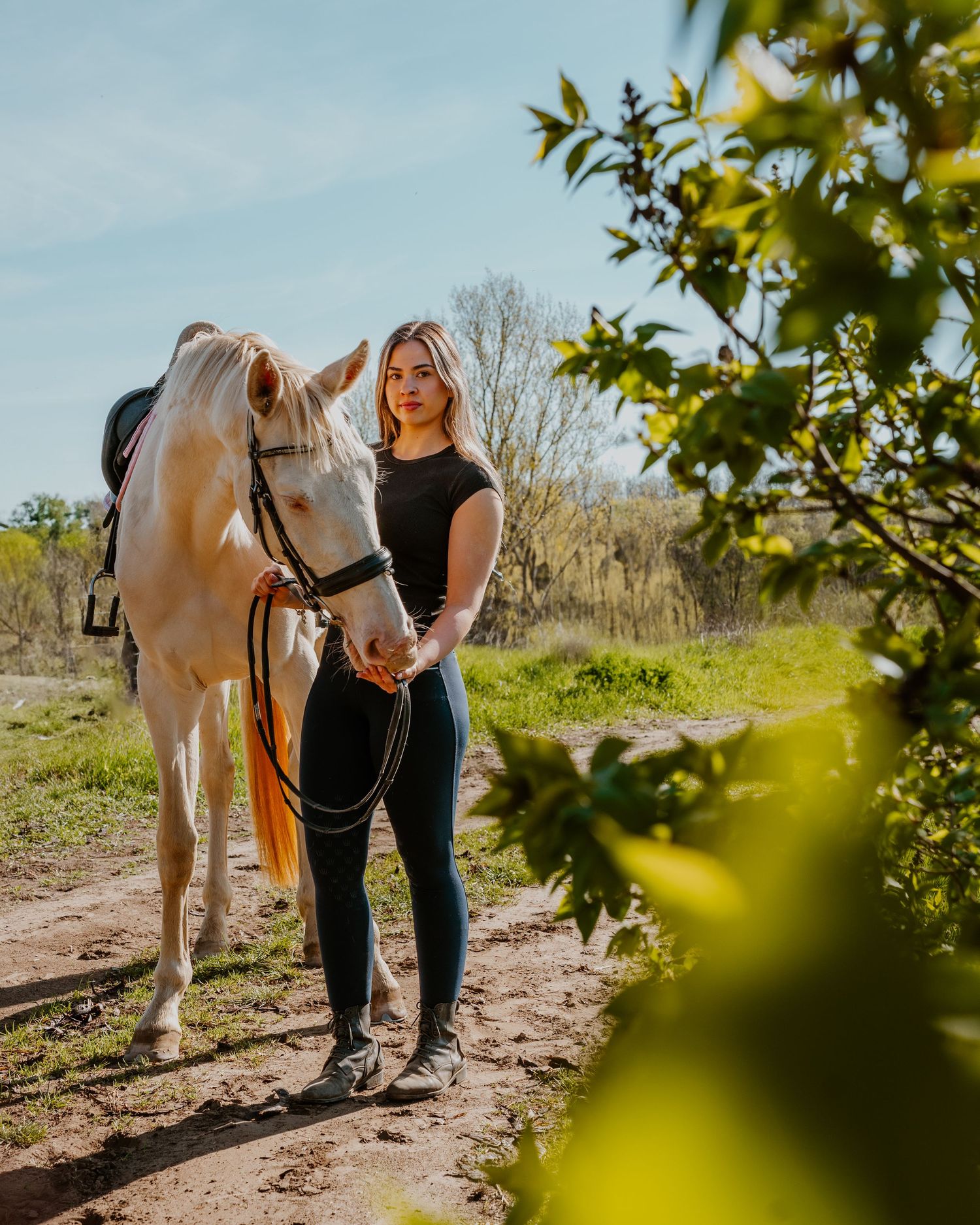 Sunny outdoor scene with someone in black standing next to a light-colored horse on a dirt path beside green foliage.