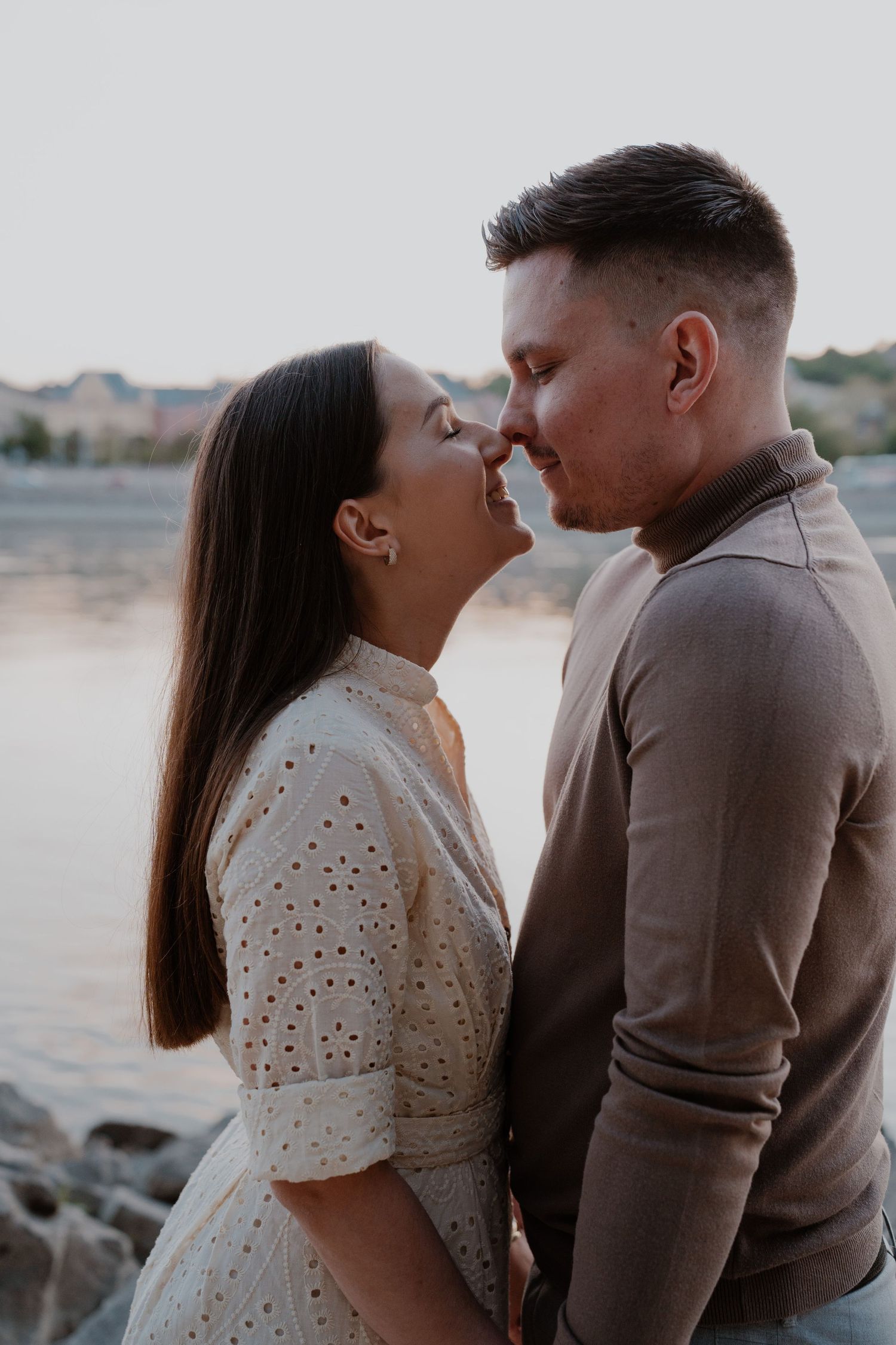 Romantic couple sharing an intimate moment near a lake at sunset, woman in white dress and man in brown shirt.