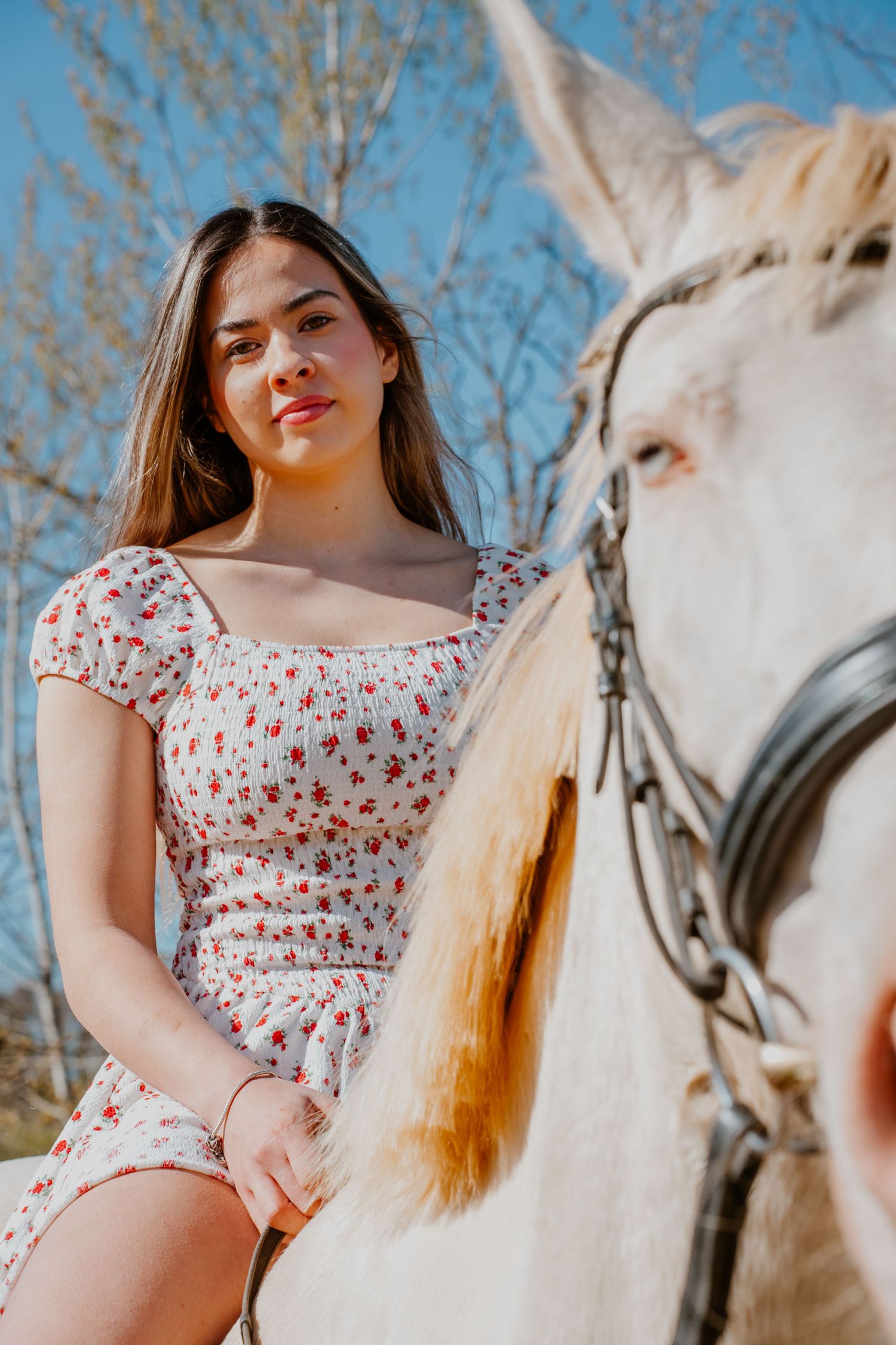 A white horse and rider in a floral print dress pose together on a sunny day against a blue sky with bare trees.
