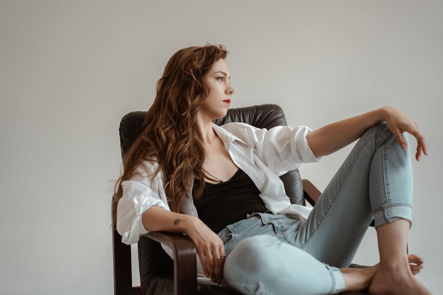 Young woman with long wavy hair sits relaxed in leather chair, wearing white shirt, black top, and light jeans.