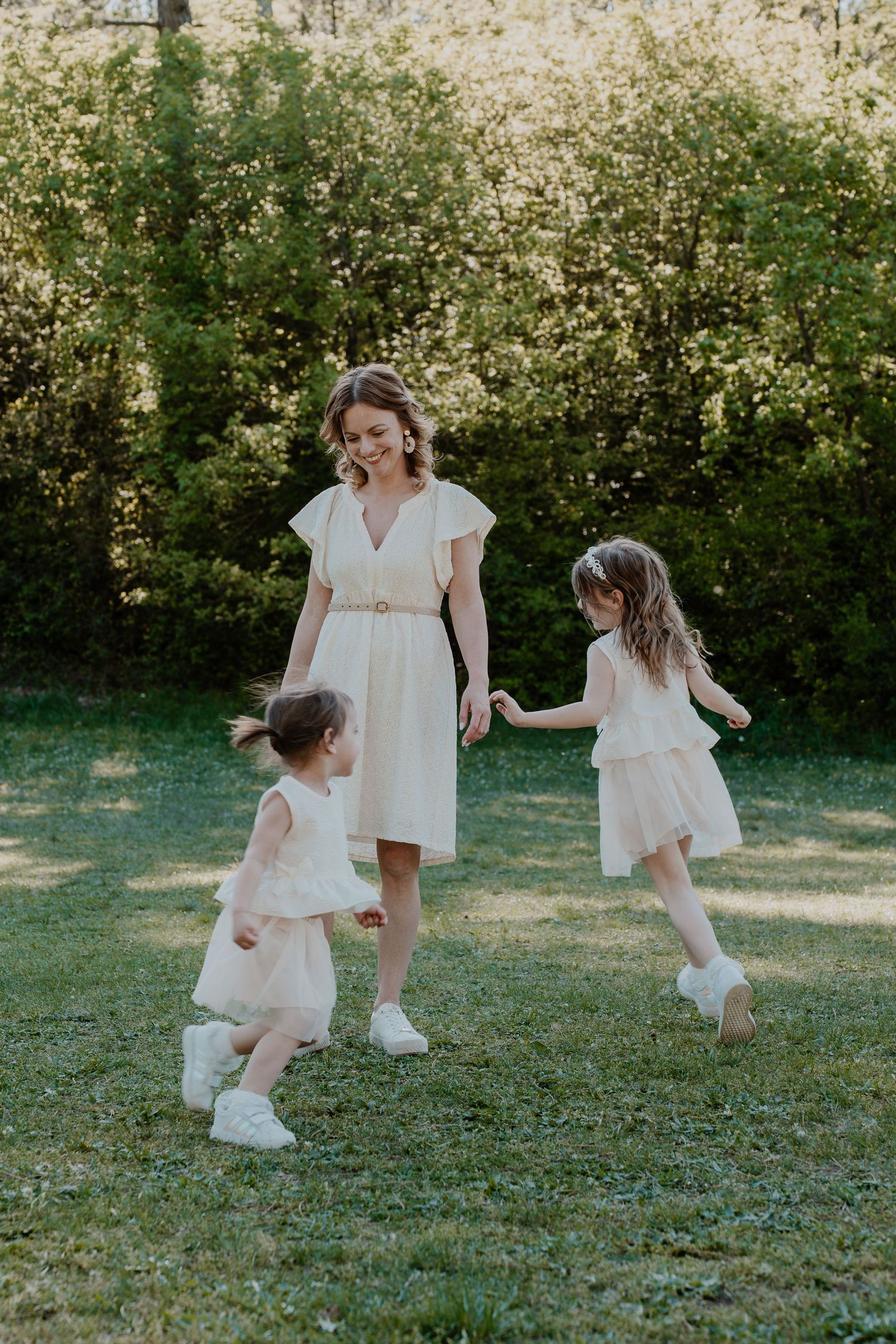 A mother in a white dress plays with her two young daughters in matching outfits on a sunny green lawn.