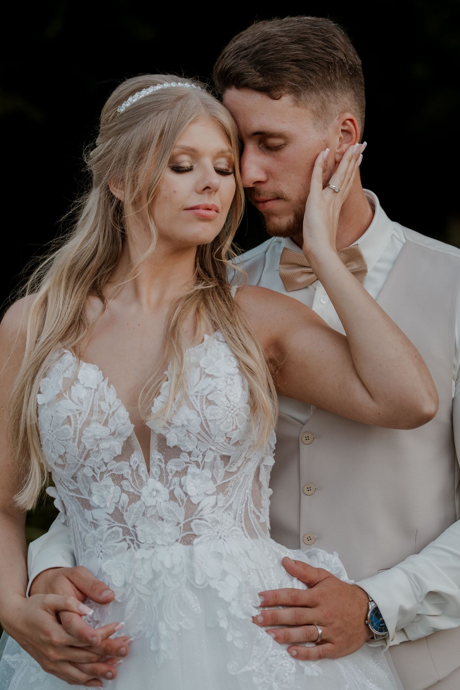 Bride in lace gown and groom in beige suit share an intimate romantic moment, eyes closed, during wedding portraits.