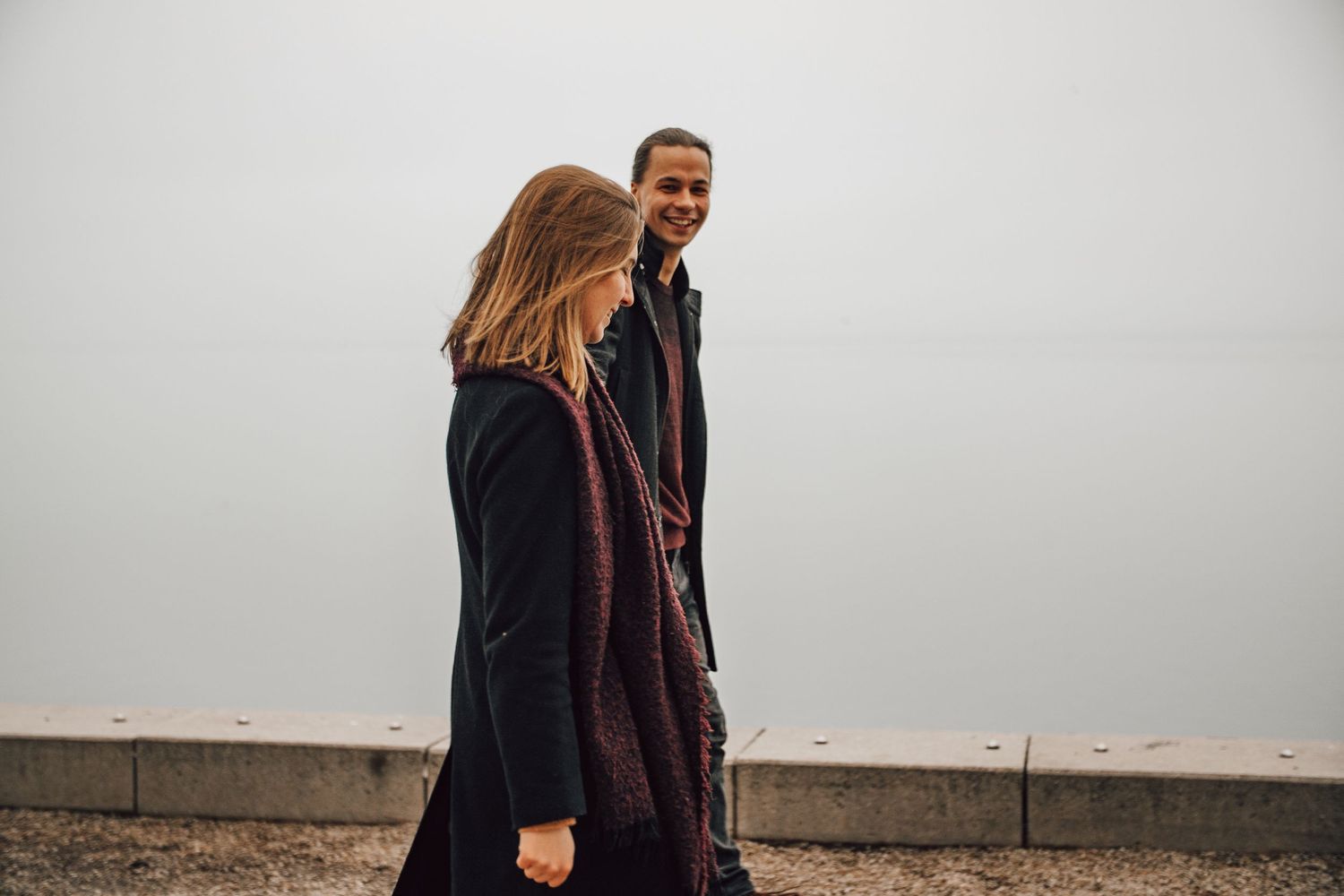 Two individuals wearing dark coats walking together on a cloudy day along a concrete surface.