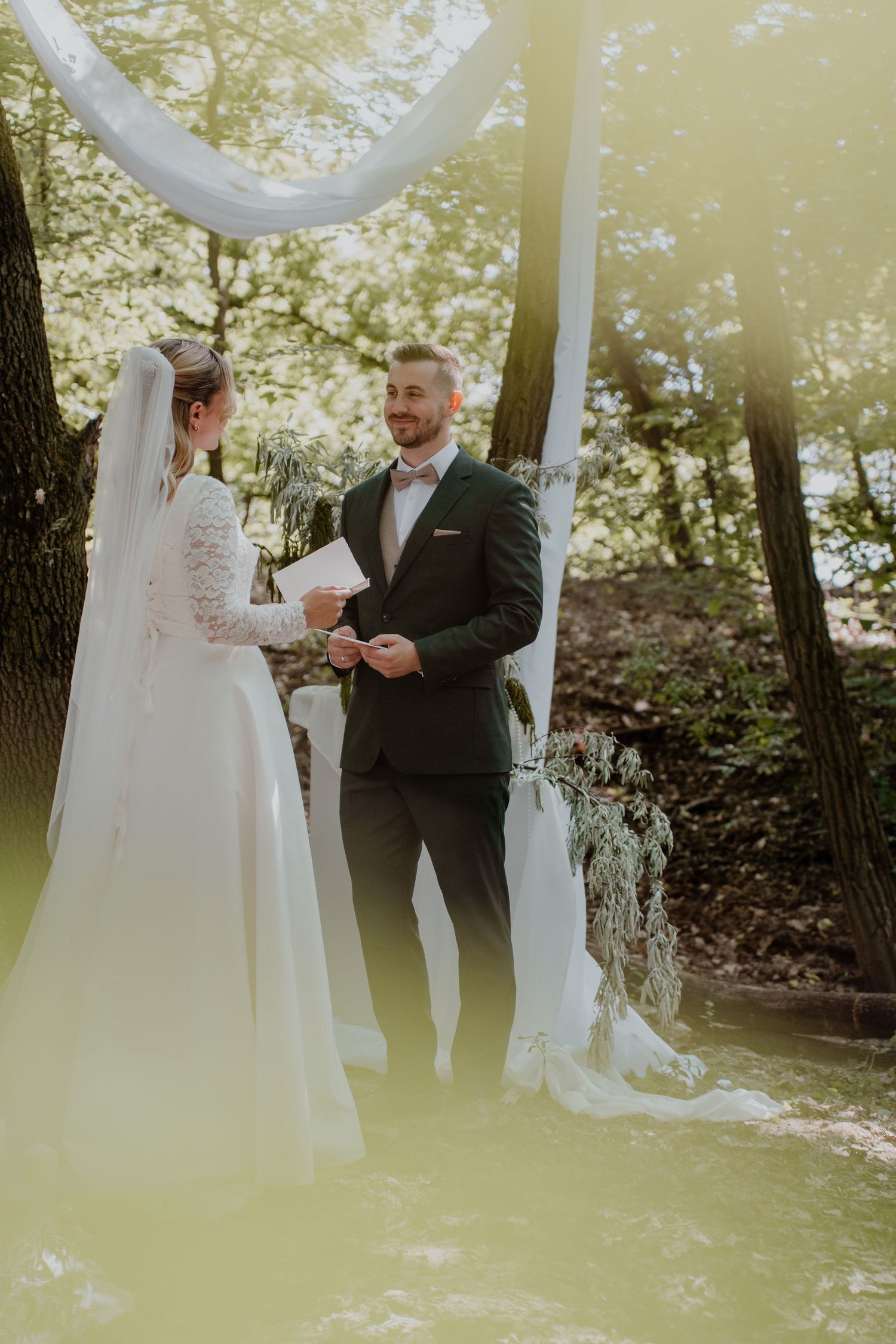 Bride and groom exchange vows outdoors in a sunlit forest wedding ceremony, with white draping and greenery in background.