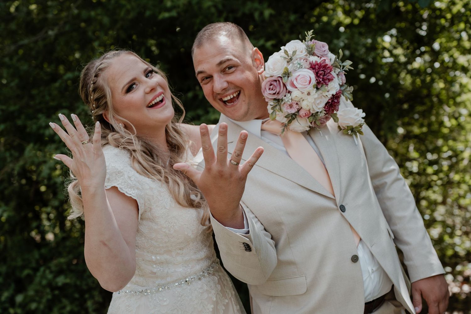 Newlywed couple joyfully showing off wedding rings, bride in white gown holding pink bouquet, groom in beige suit.