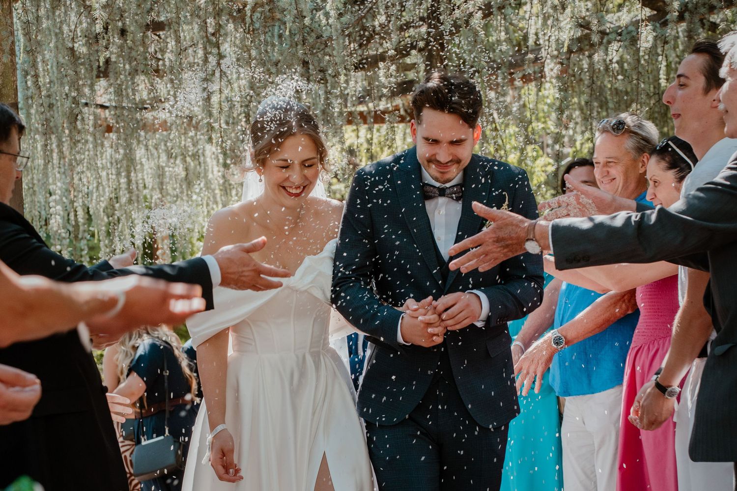 Joyful newlyweds walk through confetti shower at outdoor wedding ceremony surrounded by smiling guests.