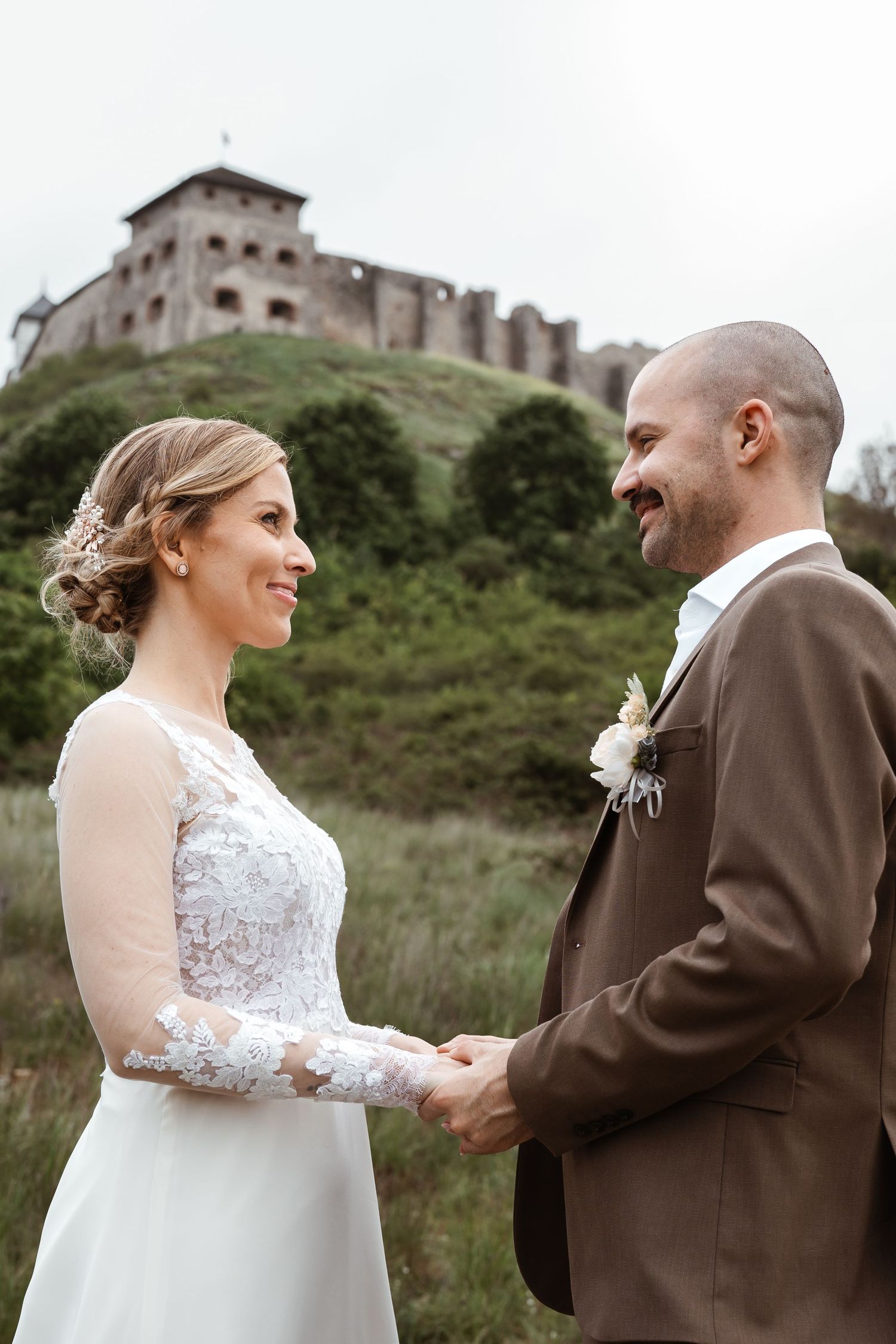 A wedding couple holds hands and smiles at each other with a historic castle perched on a hilltop in the background.