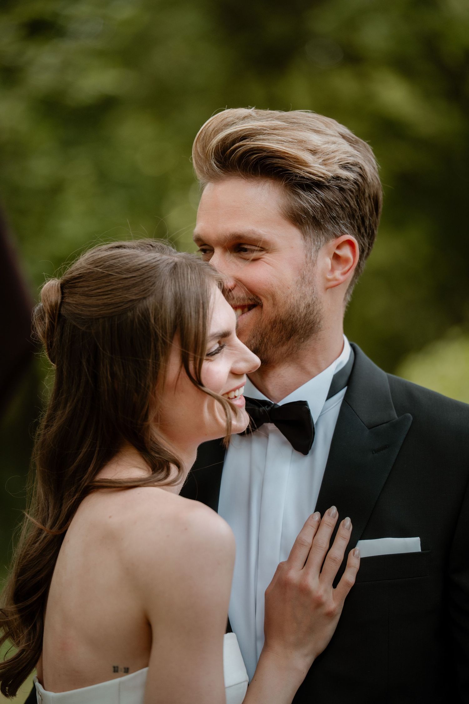 A smiling groom in a black tuxedo and bow tie leans toward his bride in a white dress outdoors.