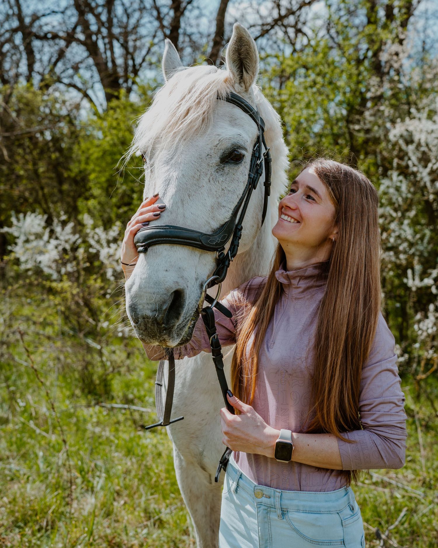 A beautiful white horse with a dark bridle stands next to someone in a pink sweater in a springtime outdoor setting.